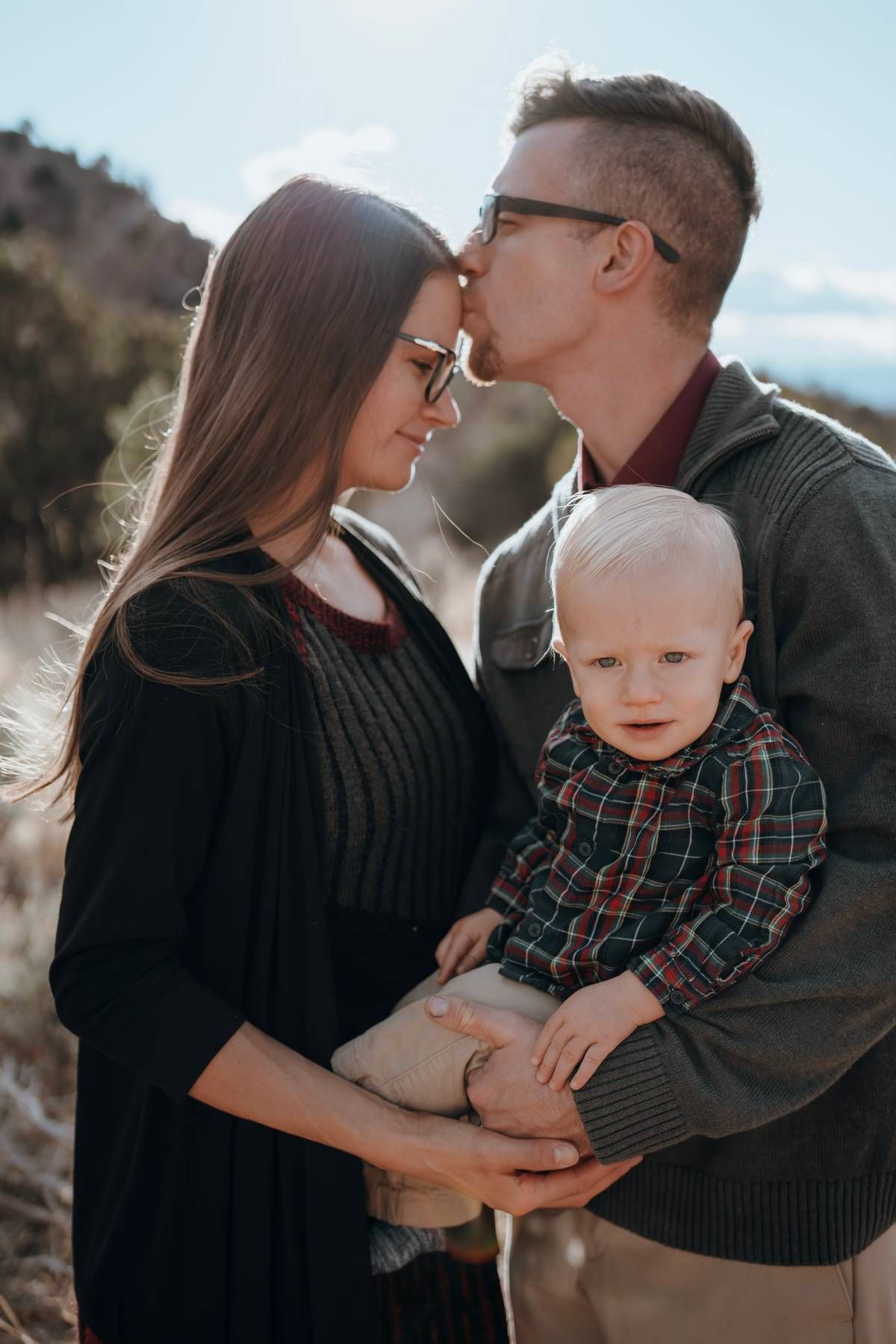 A young couple shares a kiss while holding their blonde baby boy outdoors in a scenic natural setting.