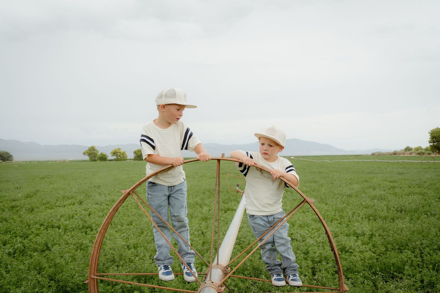 Two young boys in cowboy hats and jeans leaning on a large wagon wheel in a green farm field.