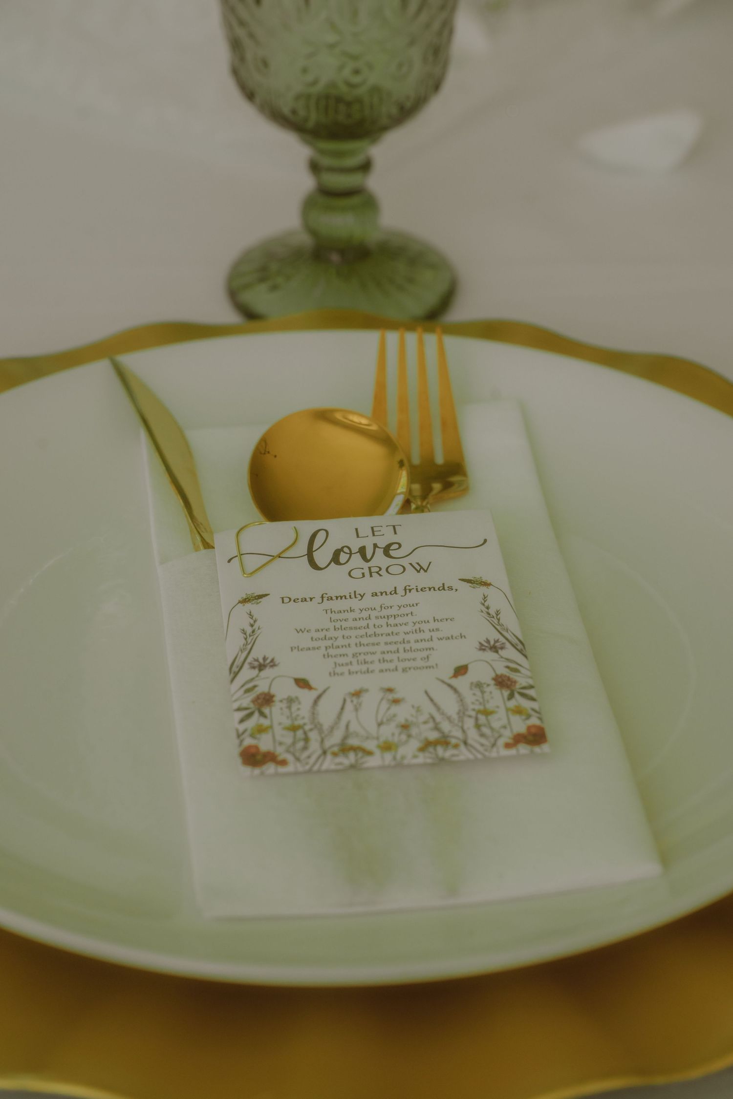 Elegant wedding place setting with gold cutlery, green goblet, white napkin, and a floral 'Let Love Grow' card on mint plate.