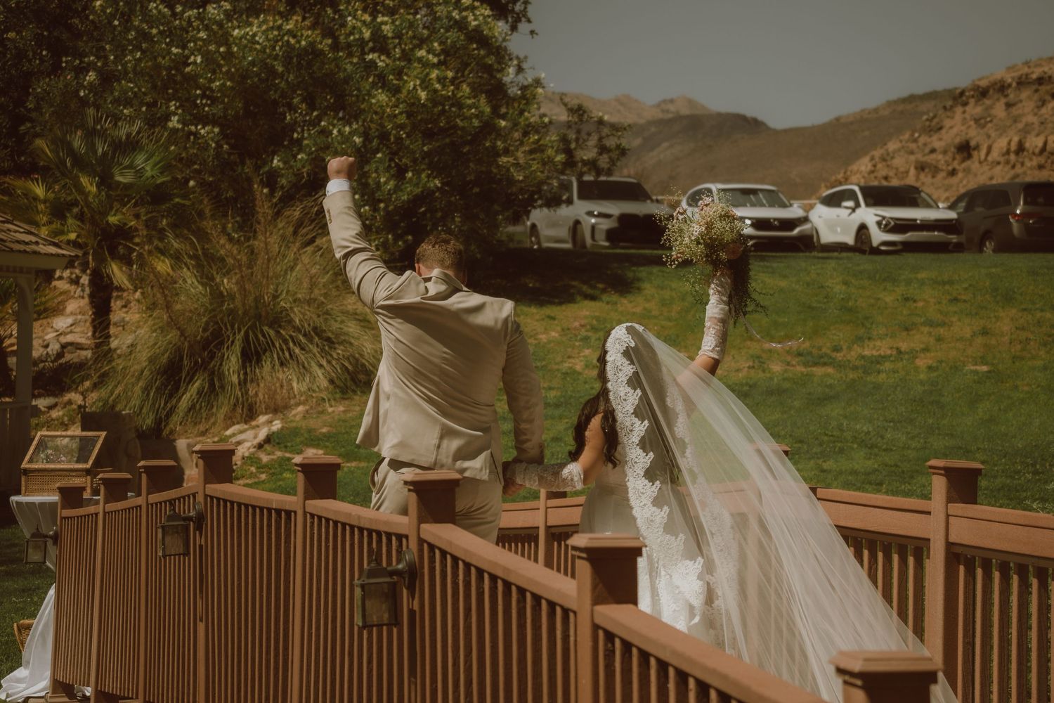 Bride and groom celebrating on a wooden deck with scenic green hills and parked cars in the background.