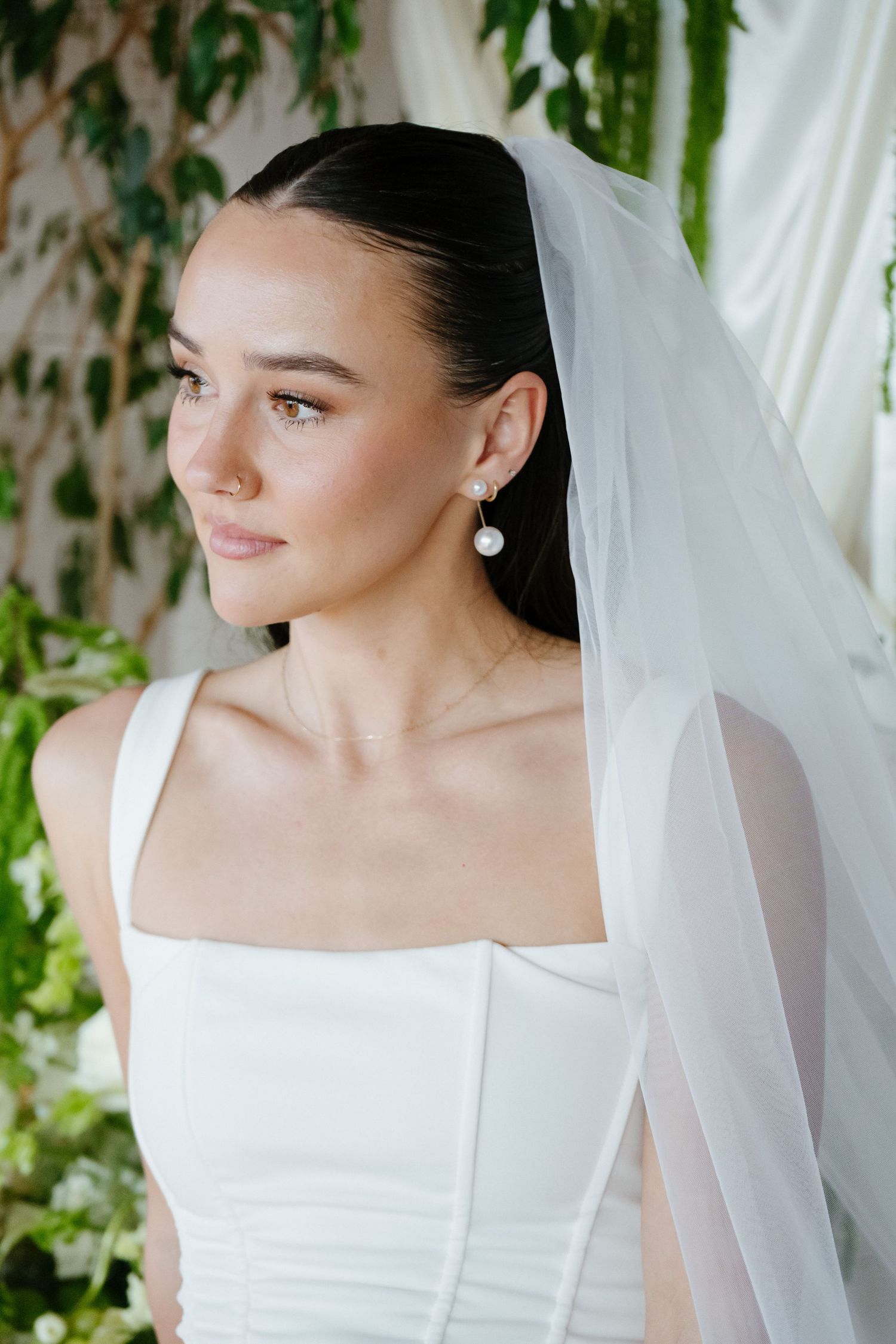 Elegant bride with dark hair in updo wearing veil and pearl drop earrings in white gown looks away.