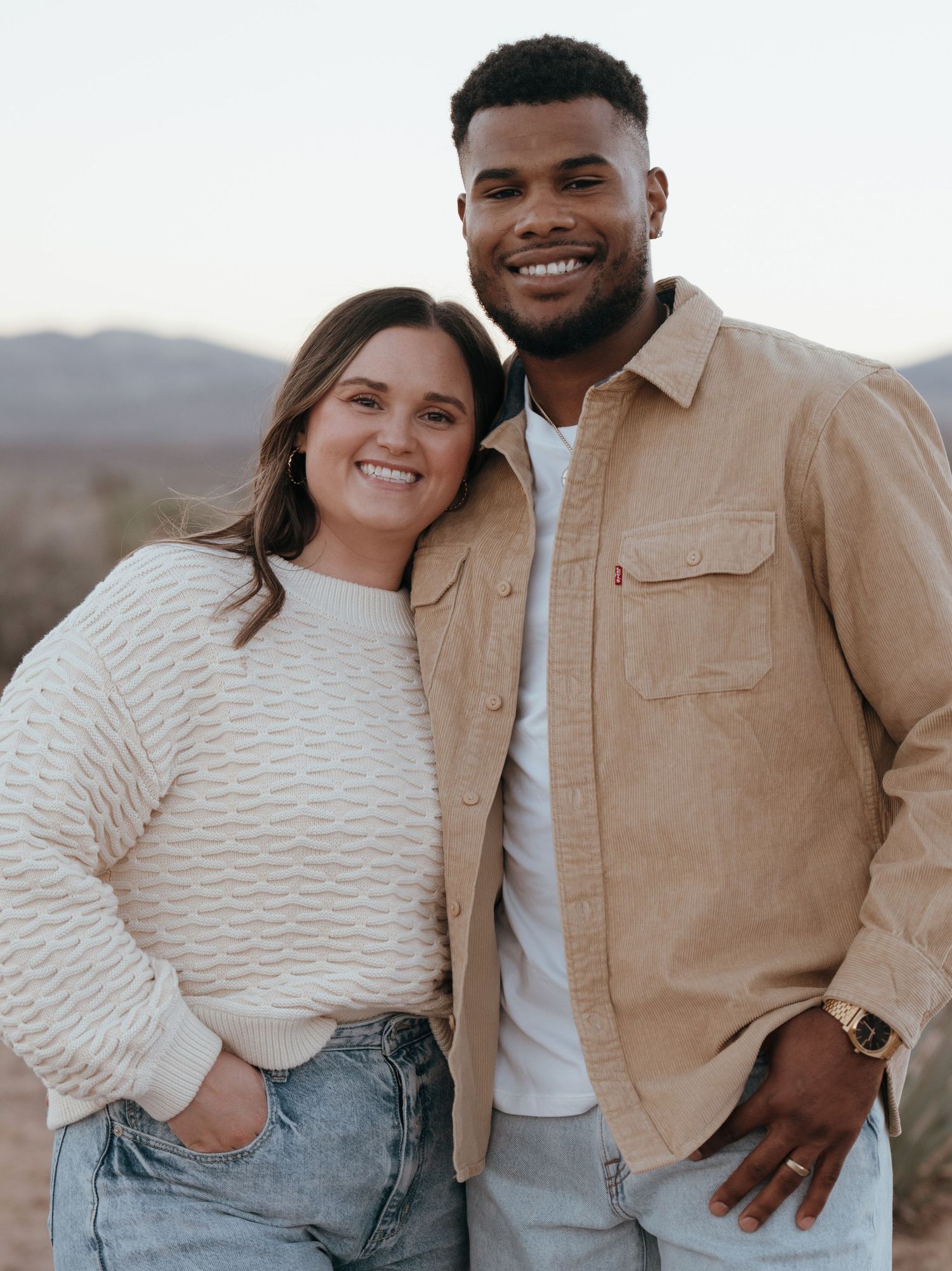 Smiling couple poses together outdoors in desert landscape at golden hour wearing casual attire.