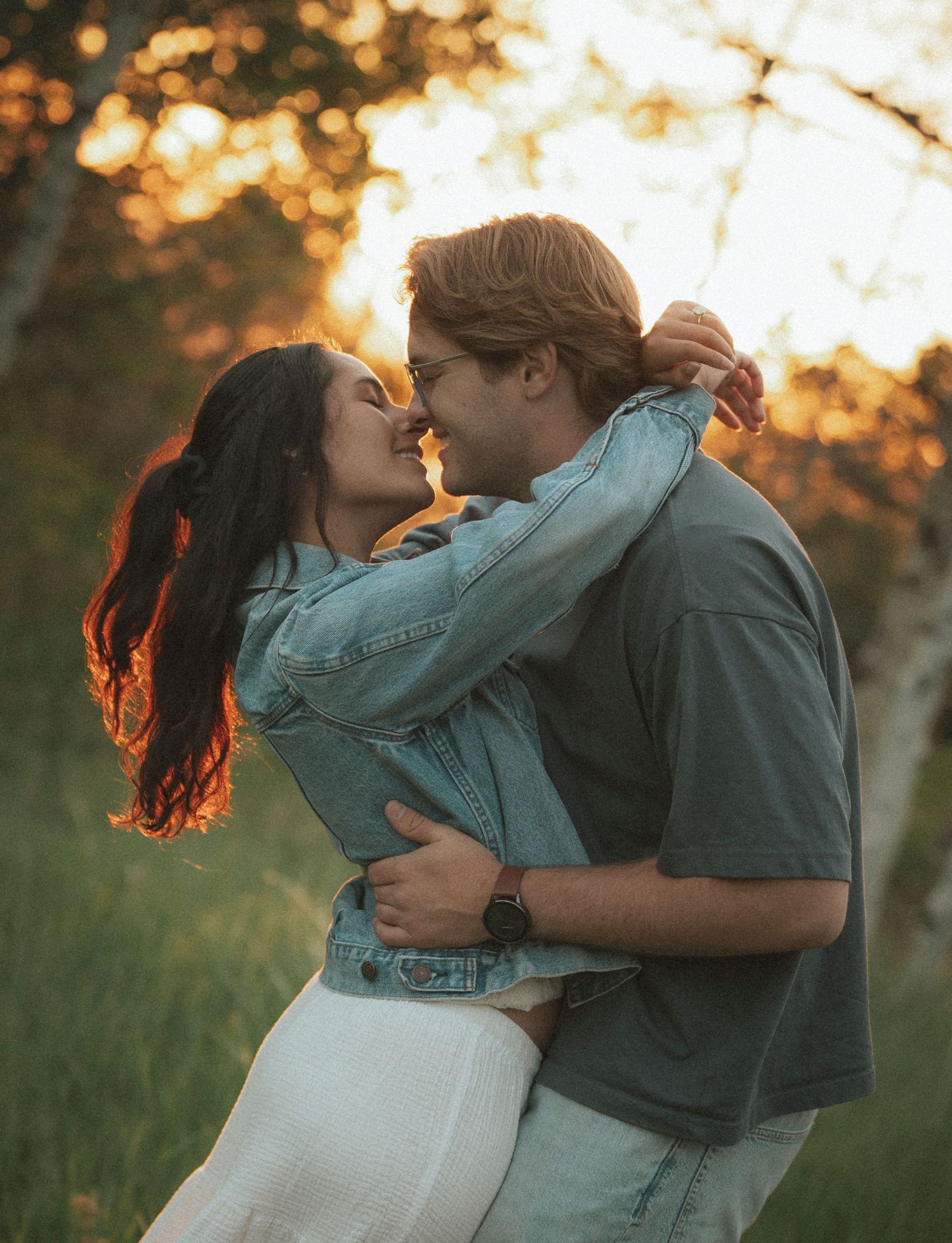 Couple embracing and almost kissing at golden hour in a green field, woman in denim jacket, man in gray shirt.