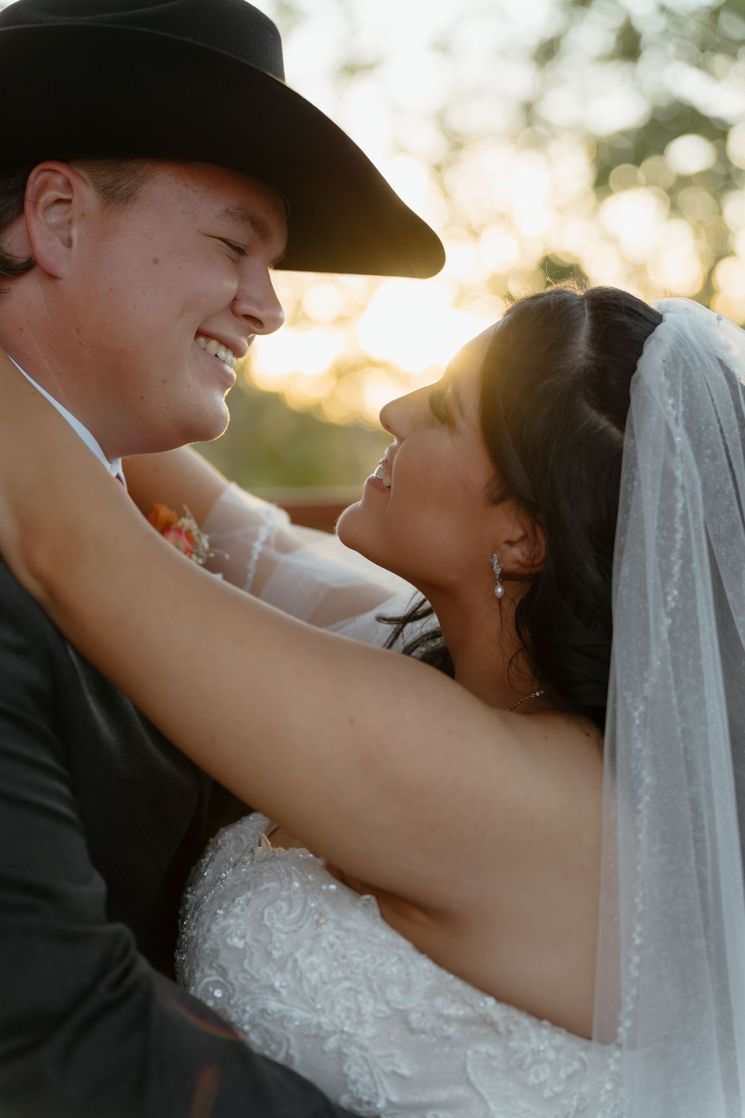 A bride and groom share a romantic moment at golden hour, with the groom wearing a black cowboy hat and the bride in a white veil.