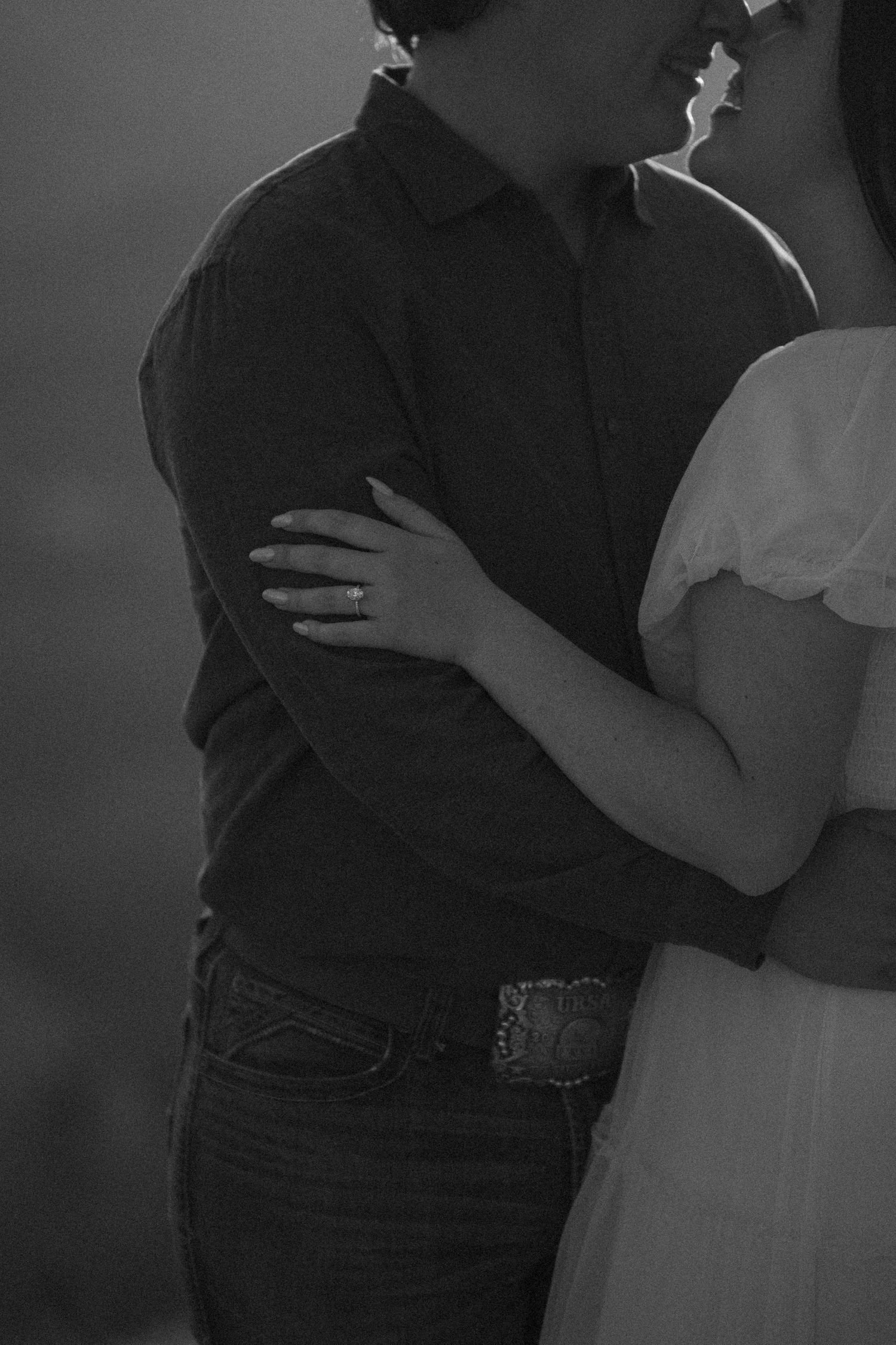 Black and white photo of a couple embracing, man in dark shirt and jeans with belt buckle, woman in white dress.