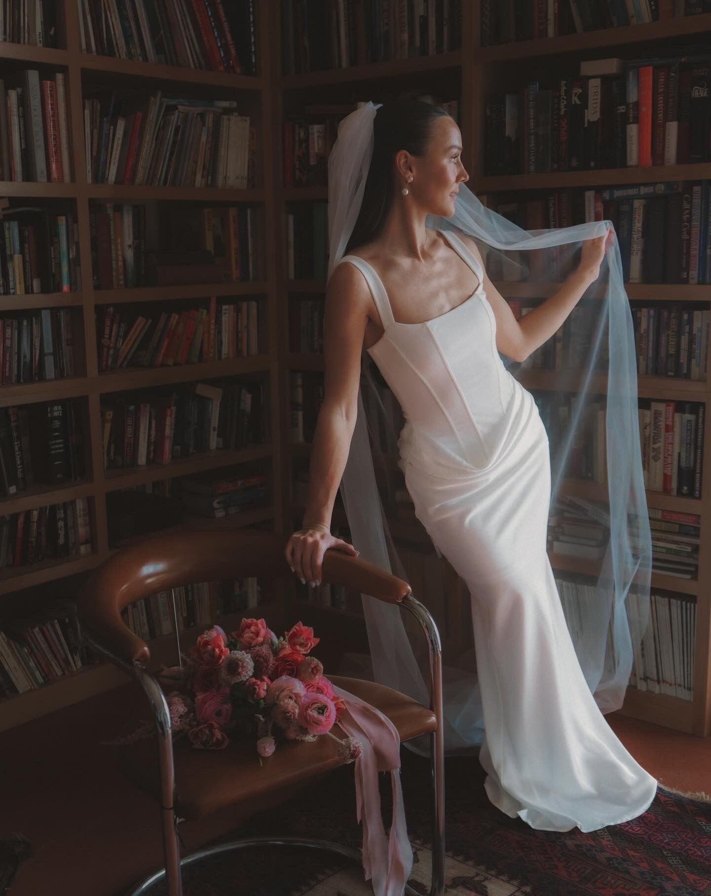 Elegant bride in a white corset gown and veil poses in a cozy library holding a pink floral bouquet.