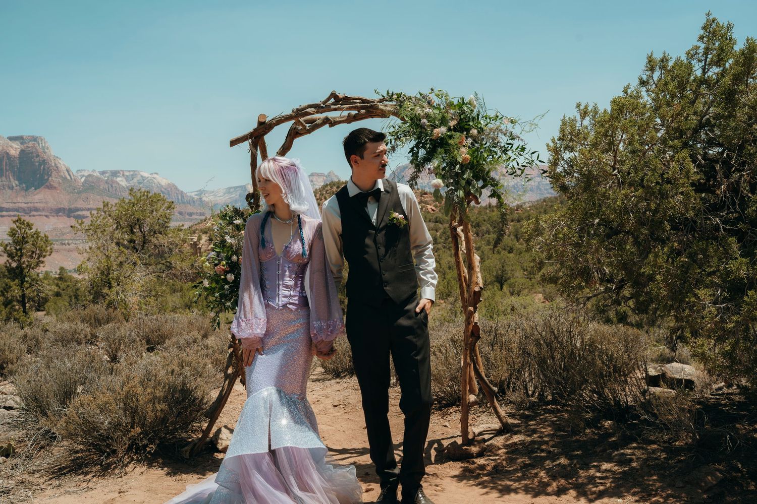 Bride and groom stand under rustic wooden arch in Sedona Arizona desert landscape with red rocks and blue sky.