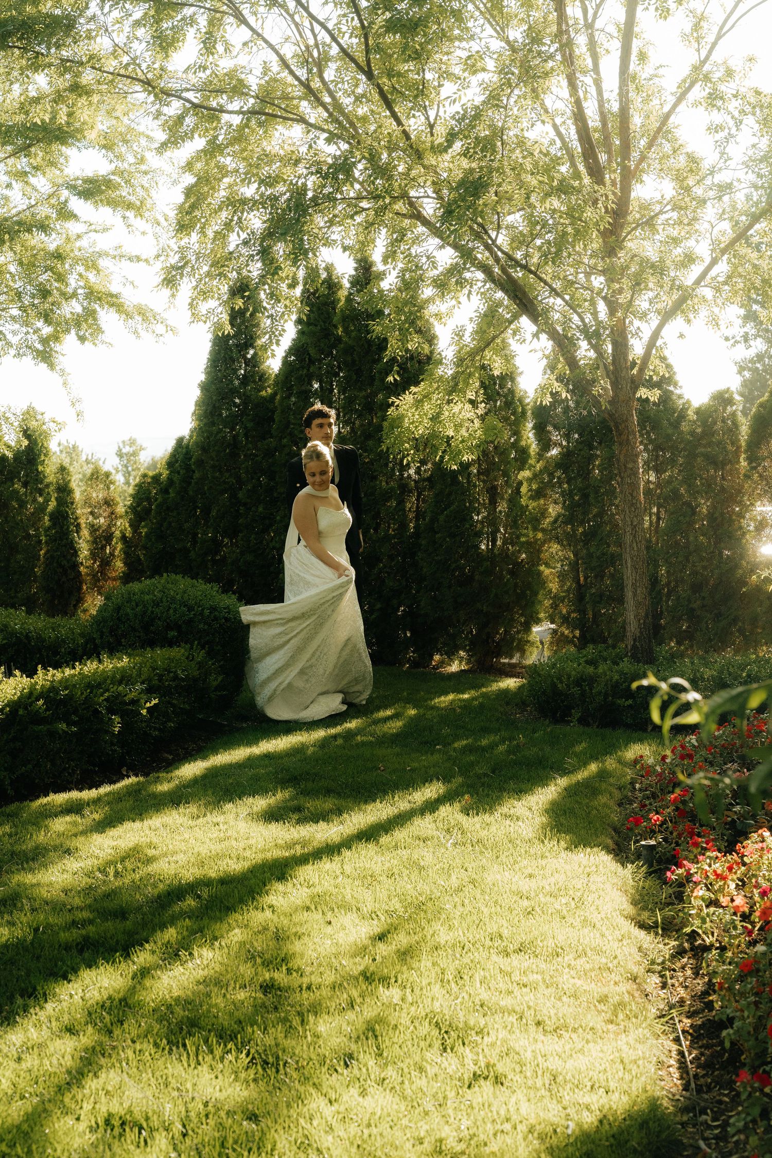 Bride in flowing white gown standing in lush garden with tall evergreens and colorful flowers in golden sunlight.