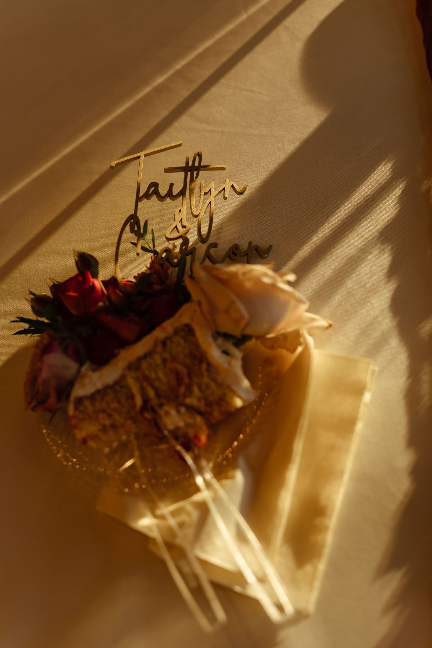 Elegant wedding cake topper with gold script names, floral decorations, and warm sunlight casting shadows.