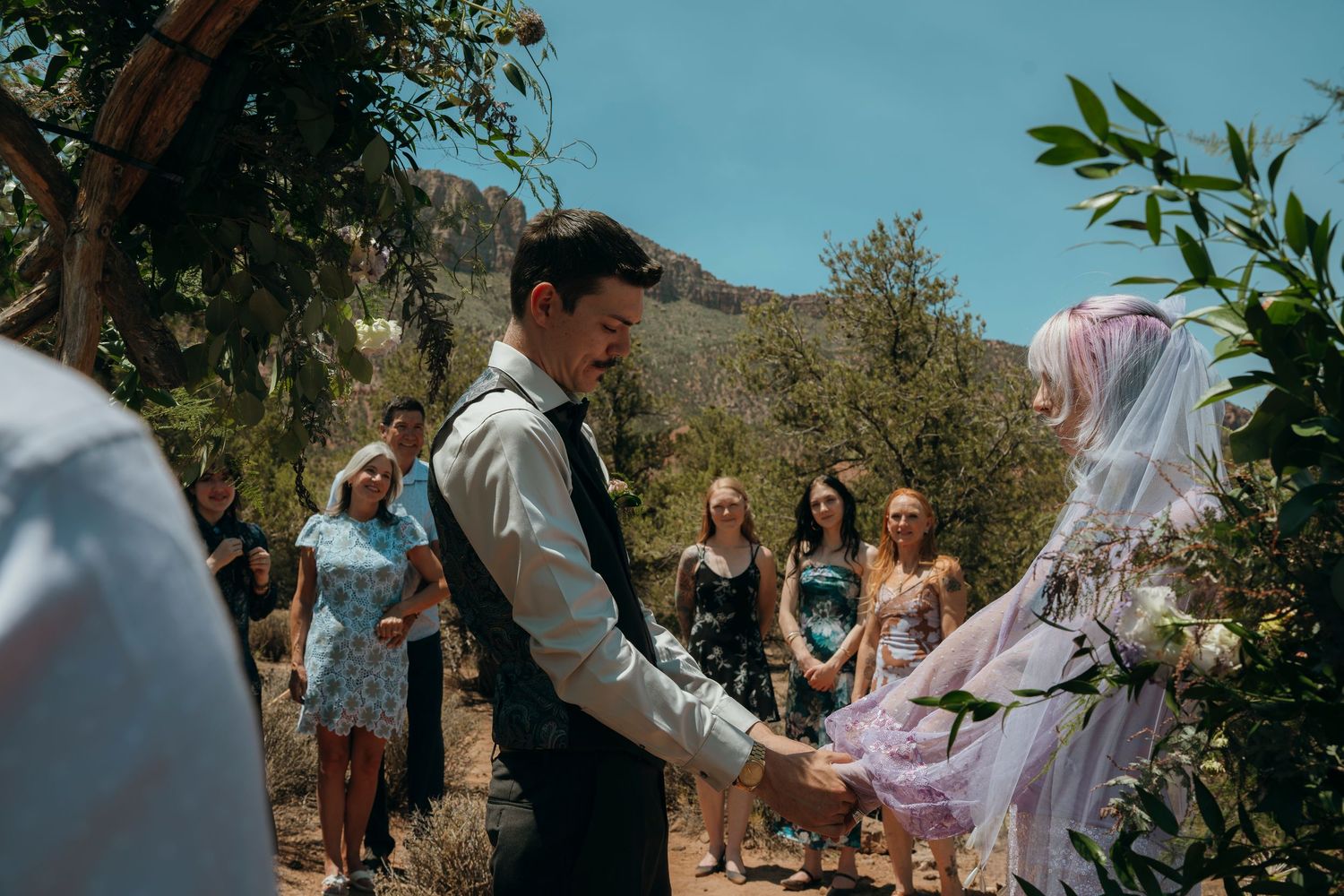 Outdoor wedding ceremony with couple holding hands, guests watching, surrounded by desert trees and mountain backdrop.