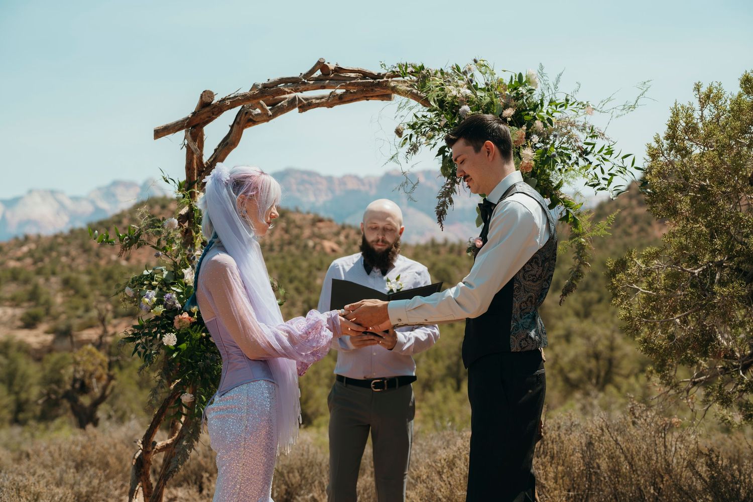 Couple exchanges vows under floral arch in desert landscape with mountain backdrop during outdoor wedding ceremony.