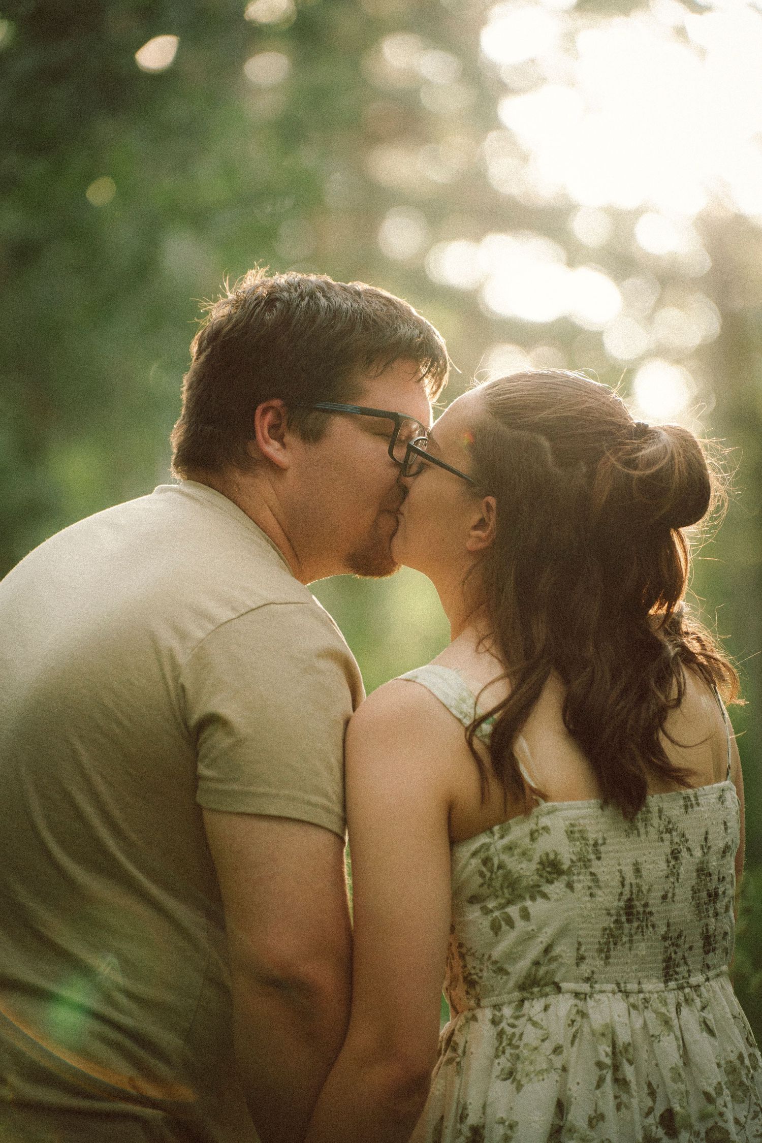 Couple sharing a romantic kiss outdoors in soft golden sunlight, both wearing glasses, surrounded by lush green trees.
