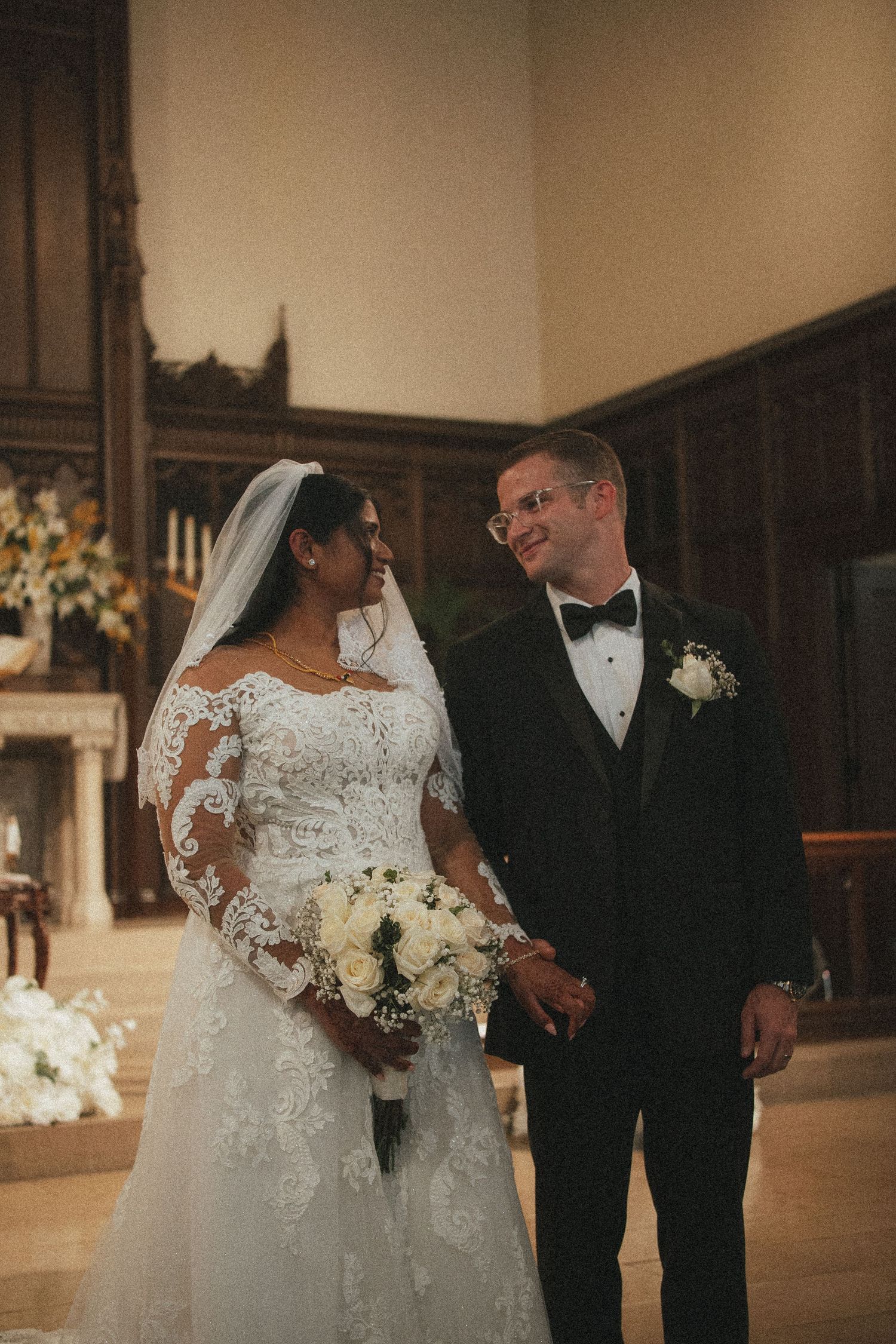 Bride in lace gown holding white bouquet stands with groom in black tuxedo at elegant church wedding ceremony.