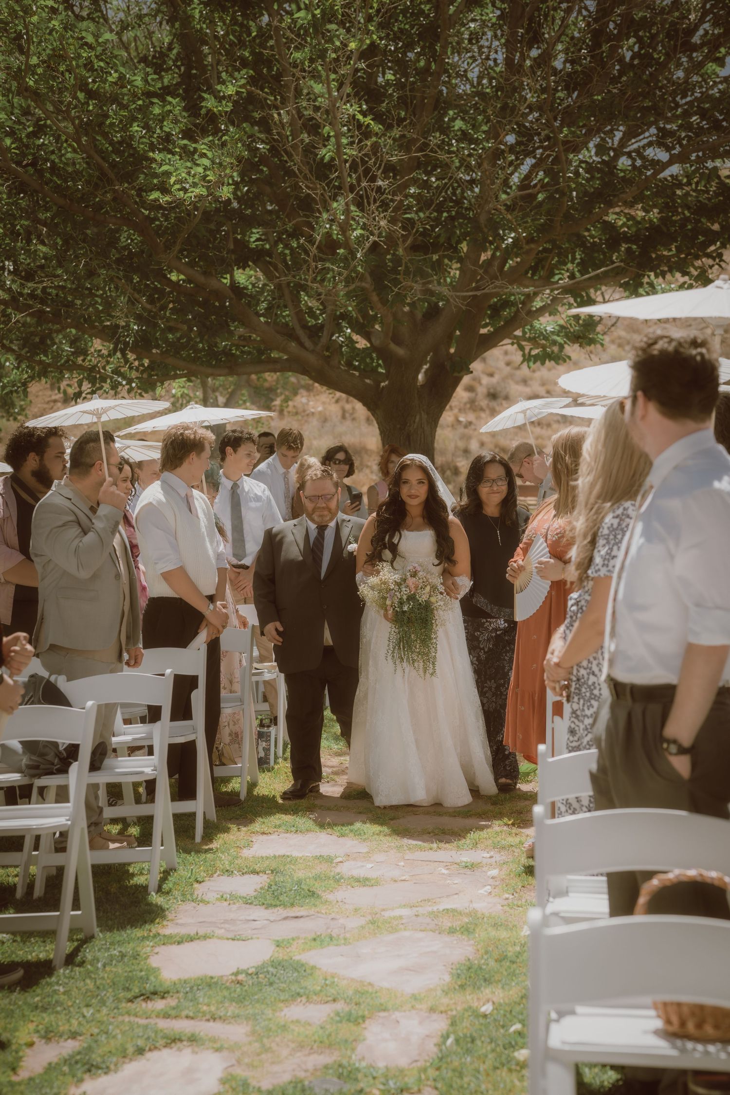 Bride walks down outdoor aisle with escort under large tree, guests standing, white chairs, holding bouquet.