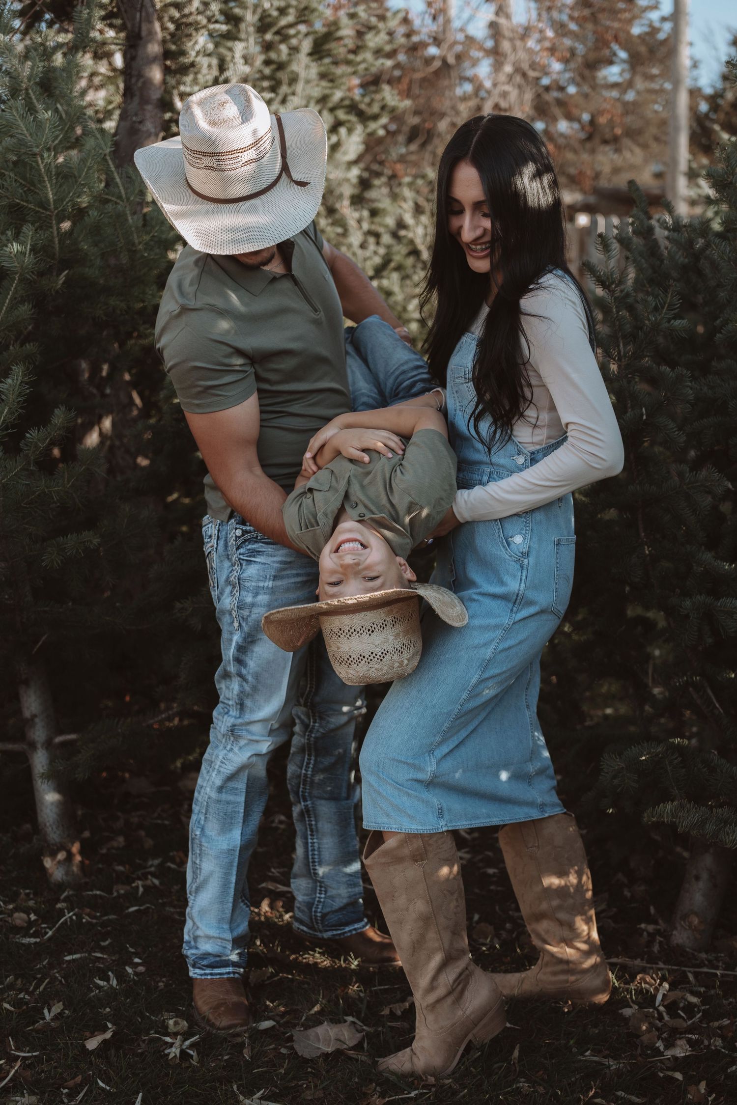 Family of three playing outdoors near pine trees, parents holding child upside down, wearing cowboy hats and denim outfits.