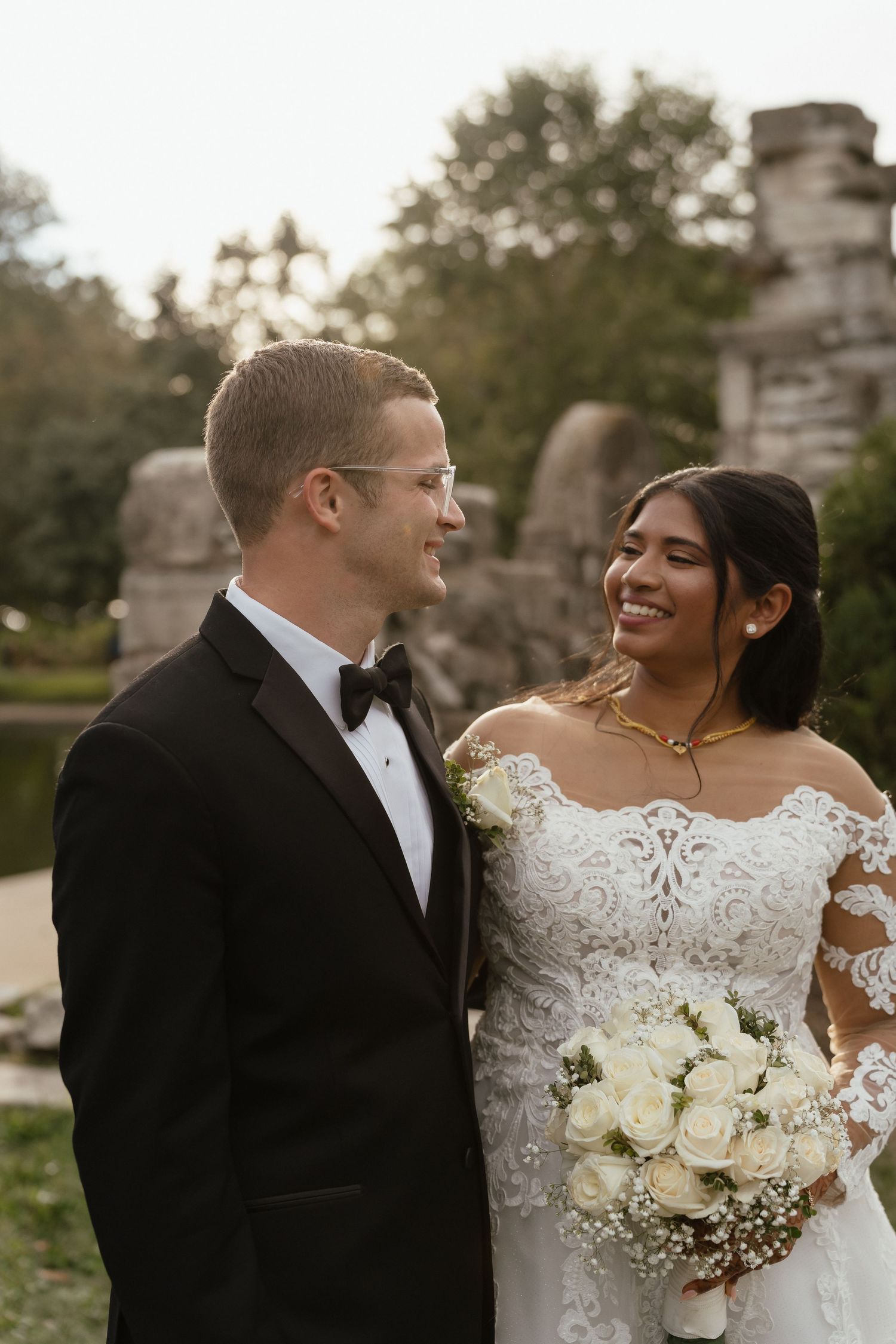 A smiling bride in lace gown holds white roses bouquet beside her groom in black tuxedo during outdoor wedding portraits.