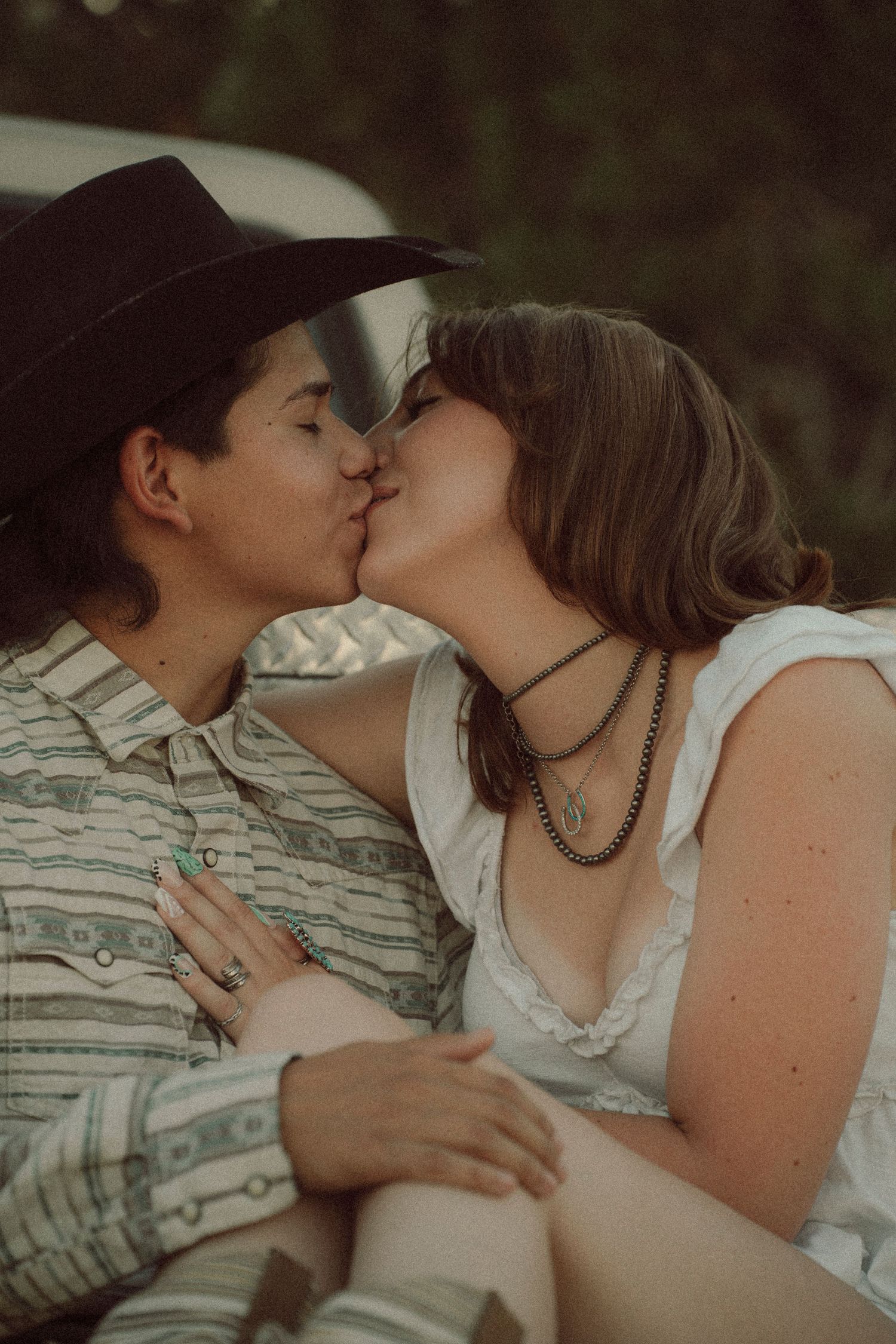 Two people sharing a romantic kiss outdoors, one wearing a cowboy hat and striped shirt, the other in a white dress.