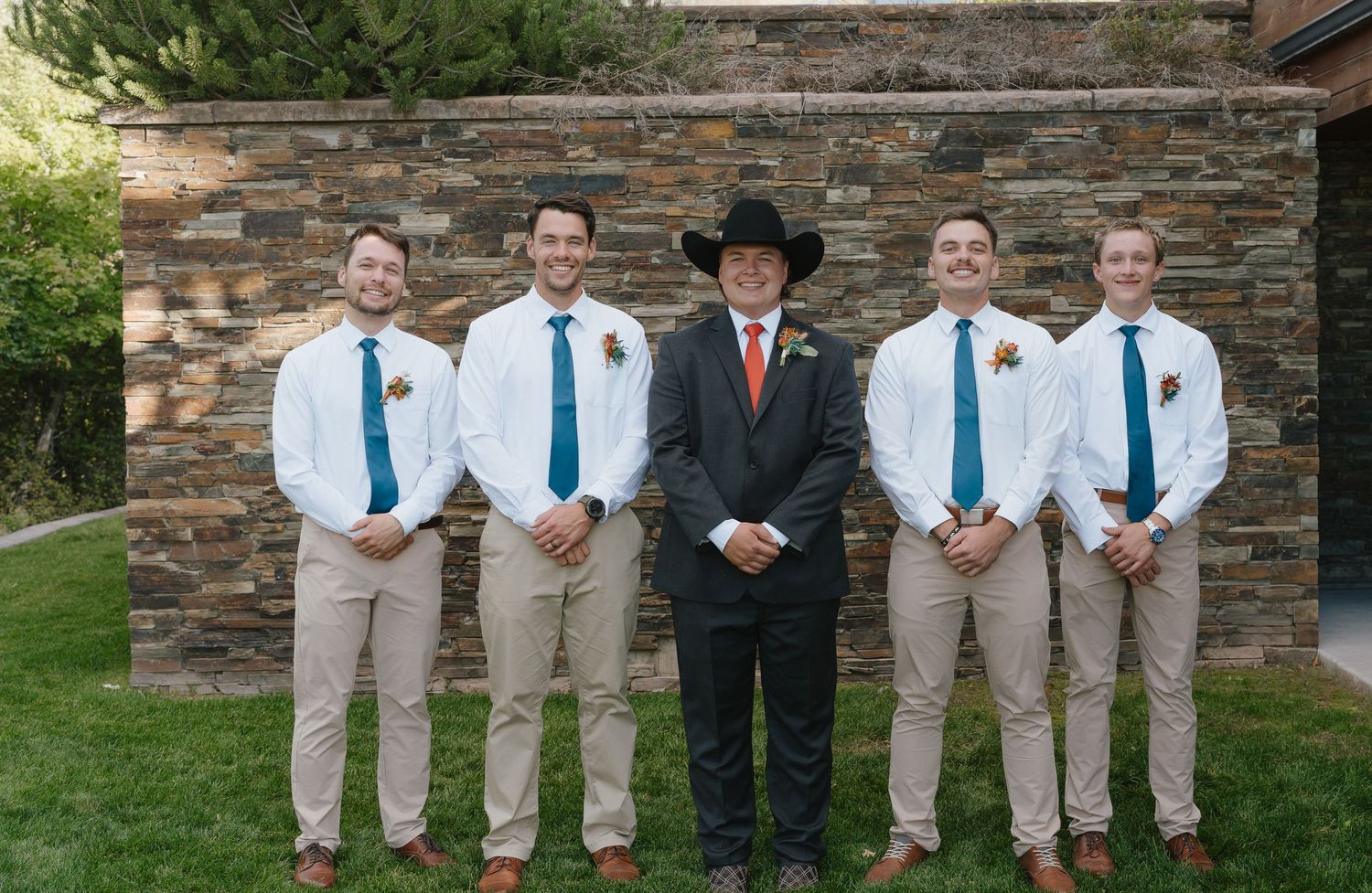 Groom in black cowboy hat and gray suit stands with four groomsmen in white shirts and teal ties outdoors.