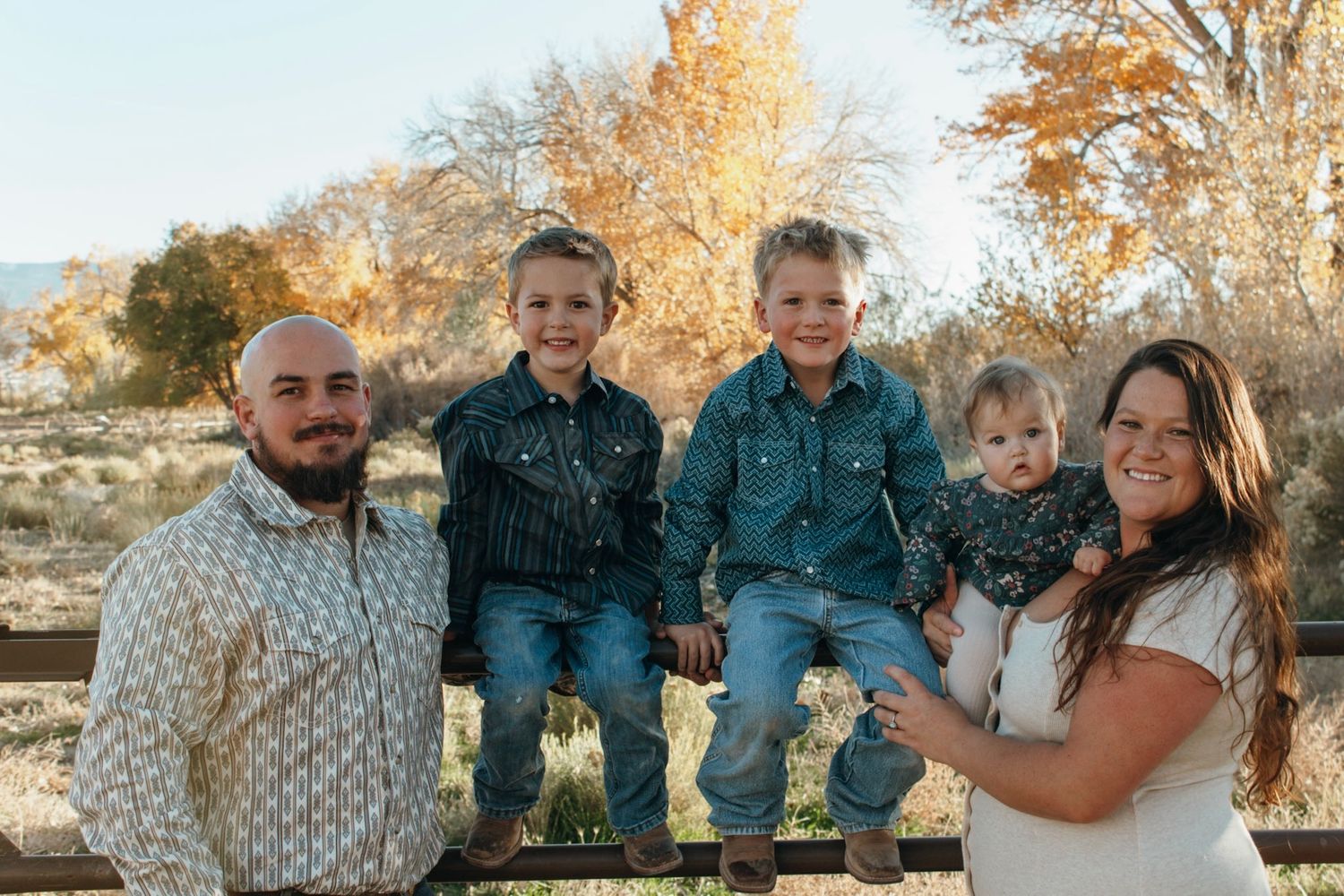 Family of five posing outdoors in fall, parents flanking three young children on a fence with autumn trees behind.