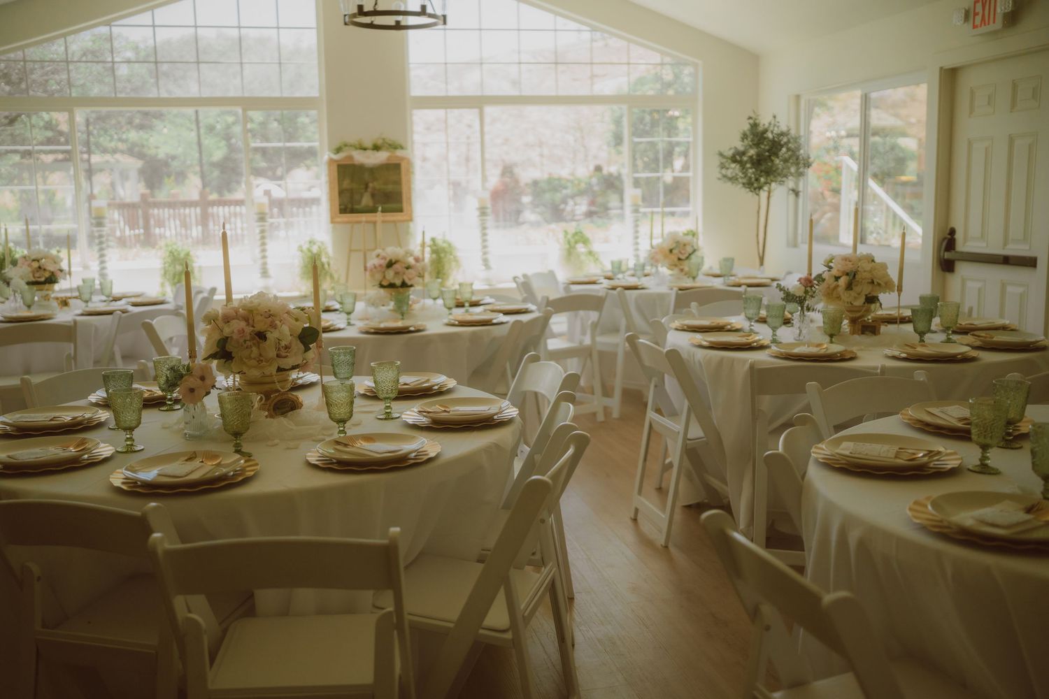 Elegant wedding reception room with round tables, white chairs, floral centerpieces, and large windows overlooking greenery.