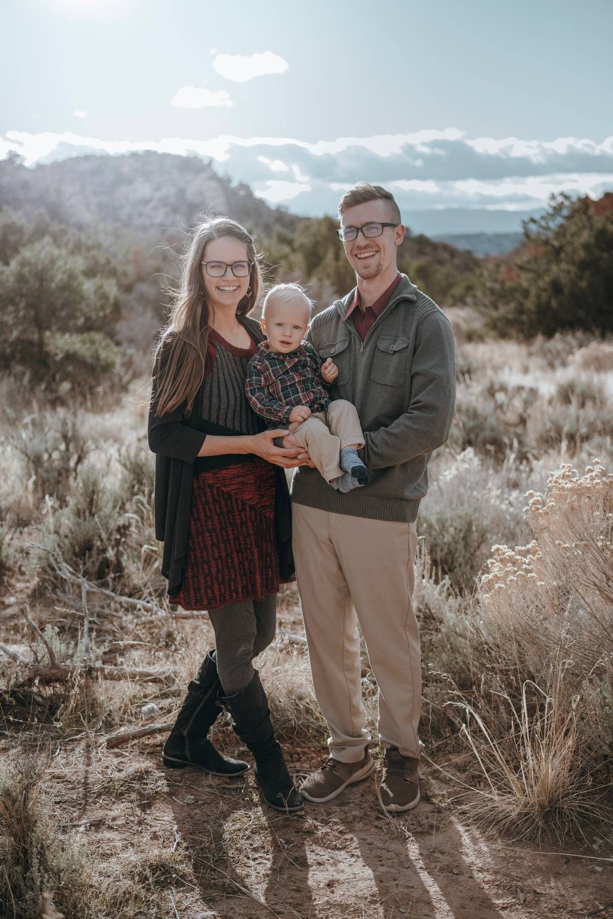 A young family of three poses outdoors in a desert landscape, with parents holding their baby on a sunny day.
