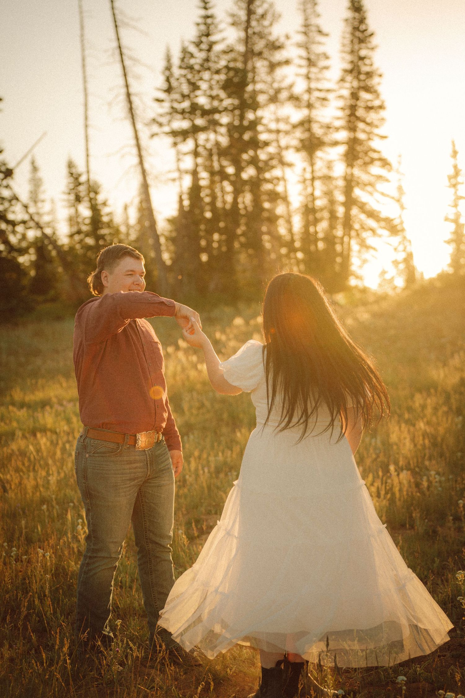 Couple dancing in golden sunset light in a meadow surrounded by pine trees, woman in white dress twirling.