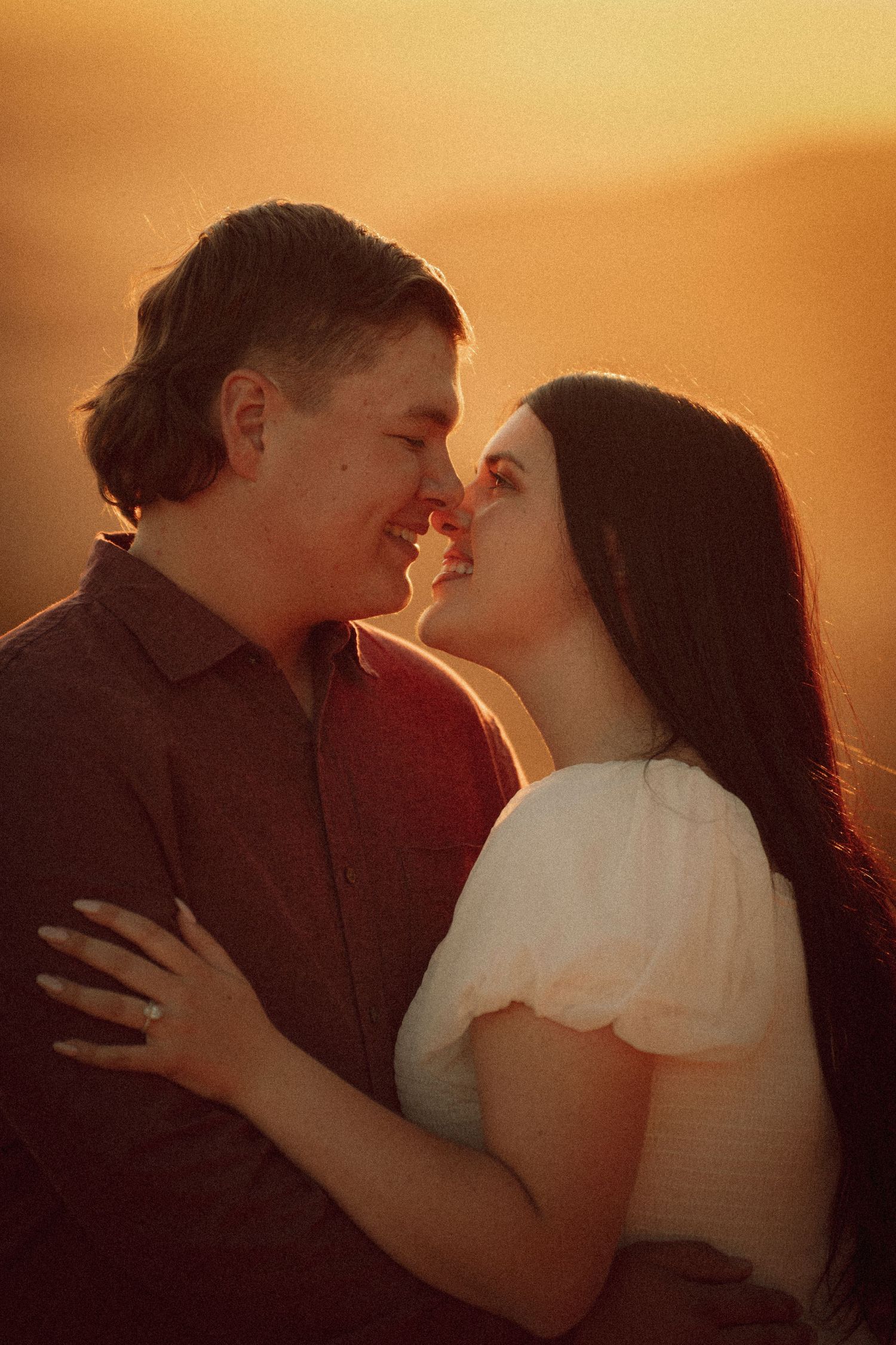 Couple sharing a romantic sunset moment, touching noses and smiling, golden hour light glowing warmly behind them.