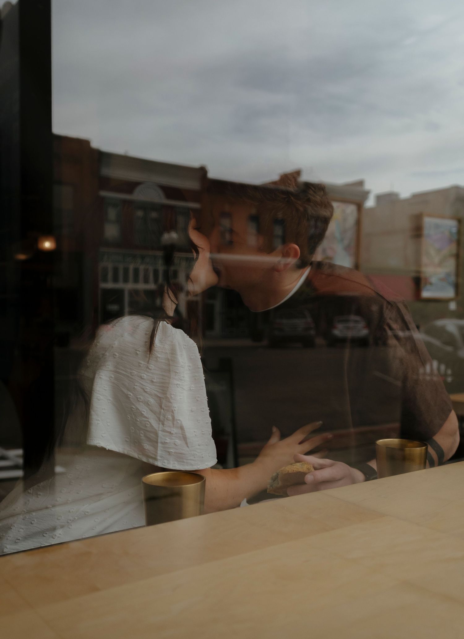 Couple sharing an intimate moment at a cafe table, viewed through a window with urban street reflection.