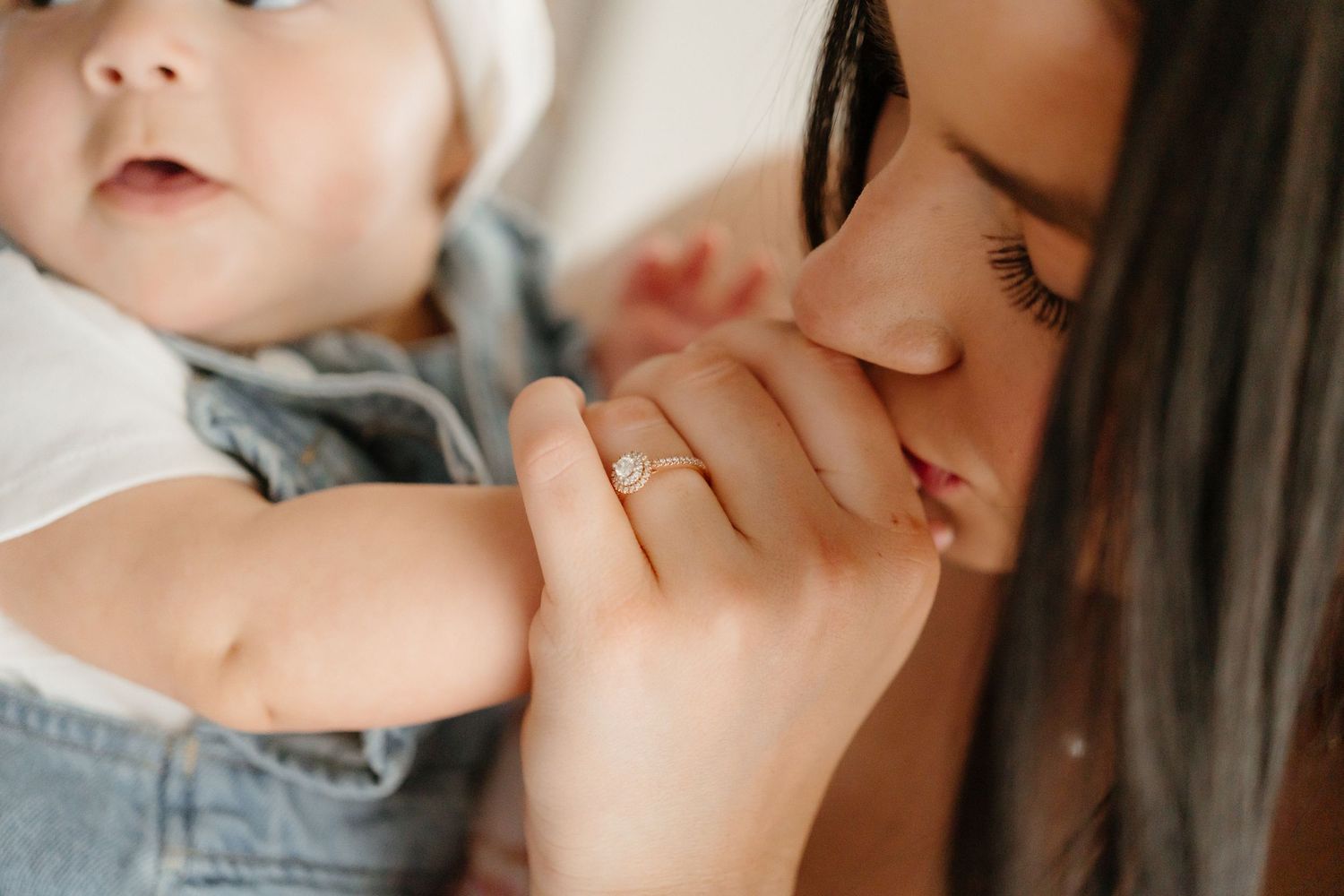Mother tenderly kissing her baby's hand while wearing a delicate diamond engagement ring.