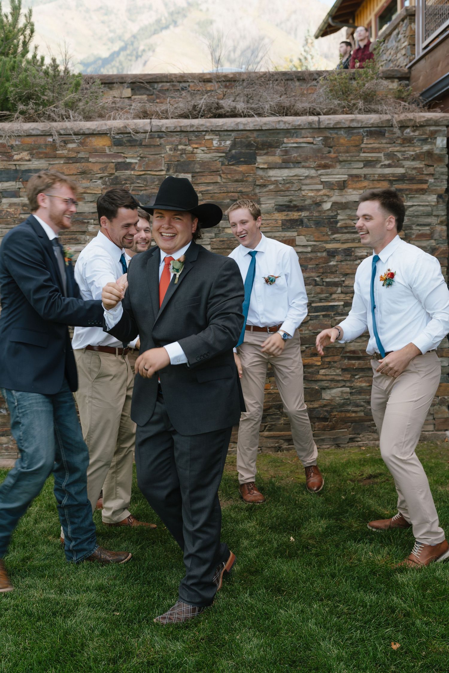 Groom in black suit and cowboy hat laughing with groomsmen in white shirts outdoors at a wedding.