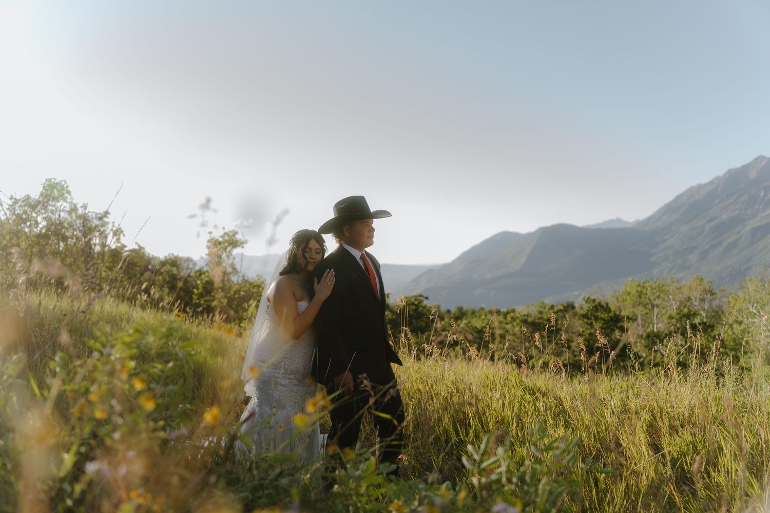 Bride and groom stand in a sunlit mountain meadow, the groom wearing a black hat, surrounded by wildflowers.