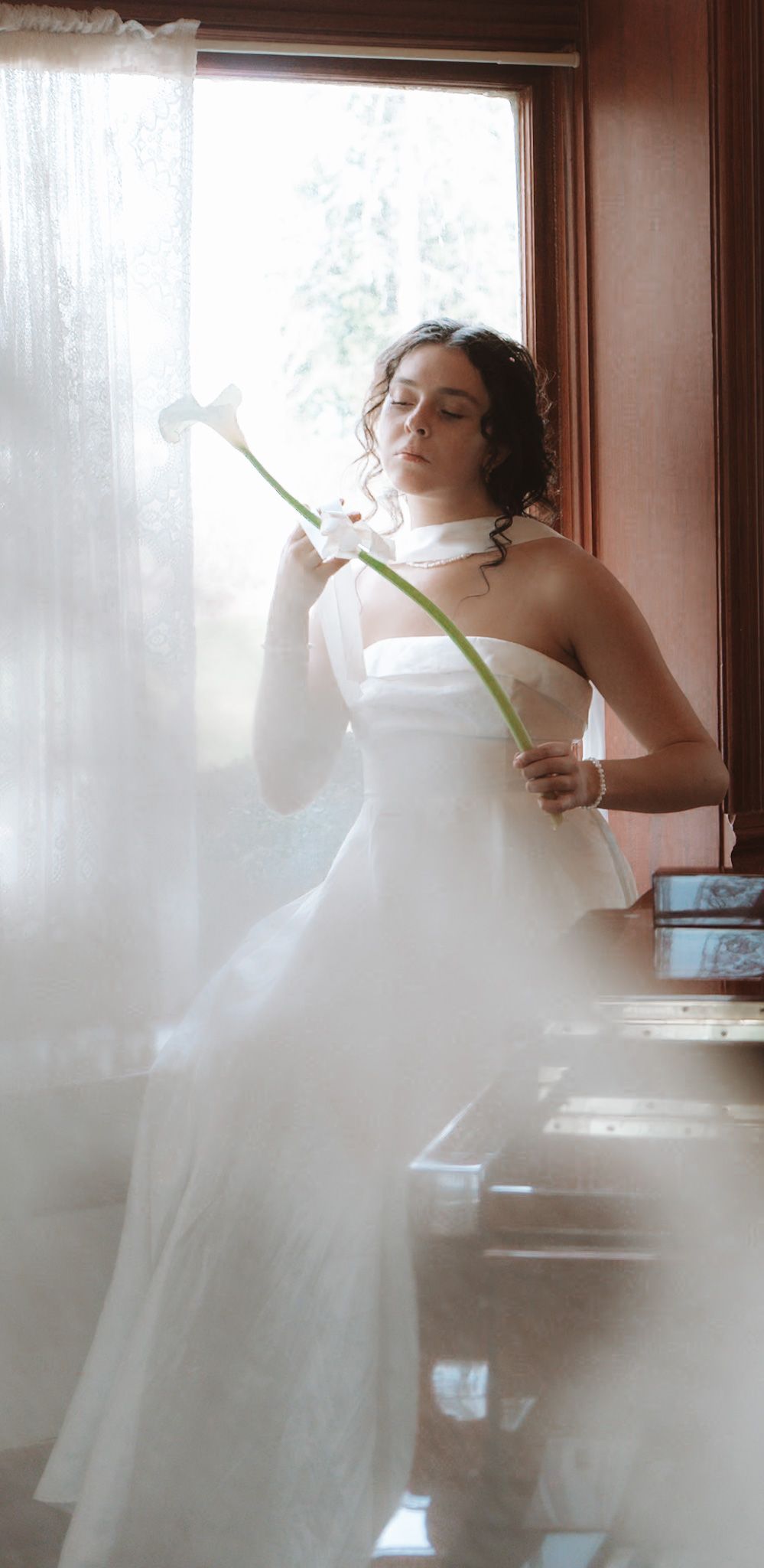 Bride in white strapless gown holding calla lilies near a sheer curtained window by a piano.
