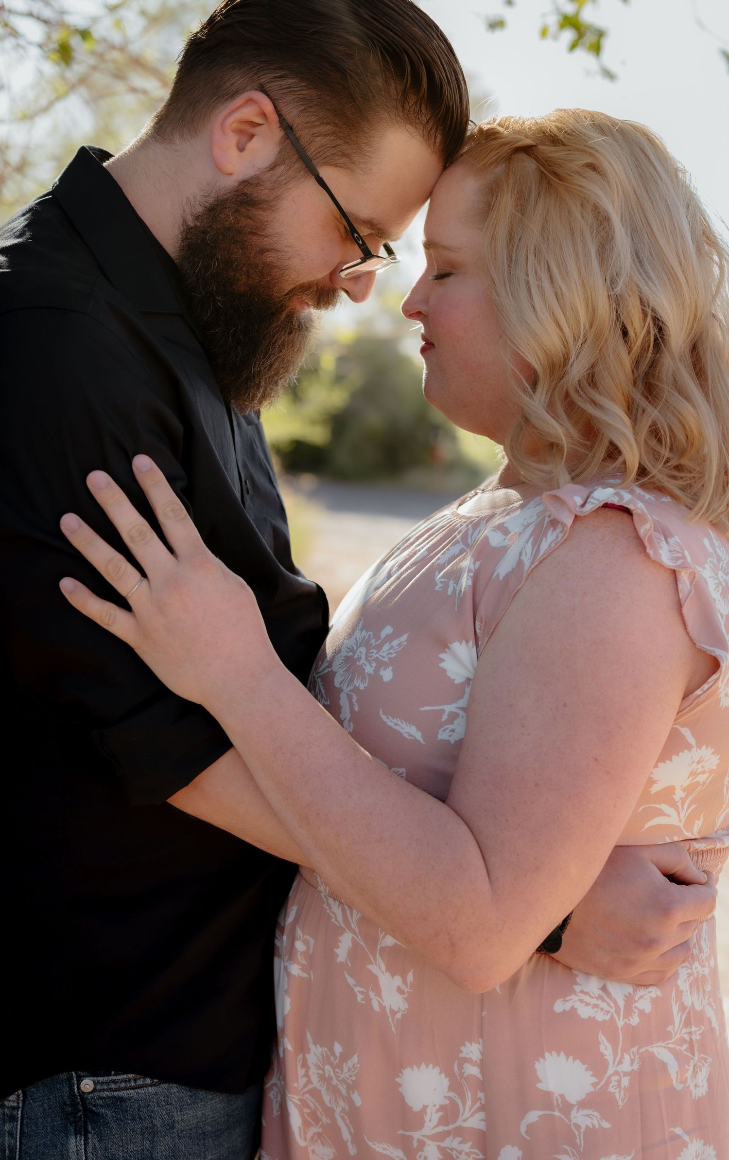 A couple shares a romantic forehead-to-forehead moment outdoors, man in black shirt and woman in floral pink dress.