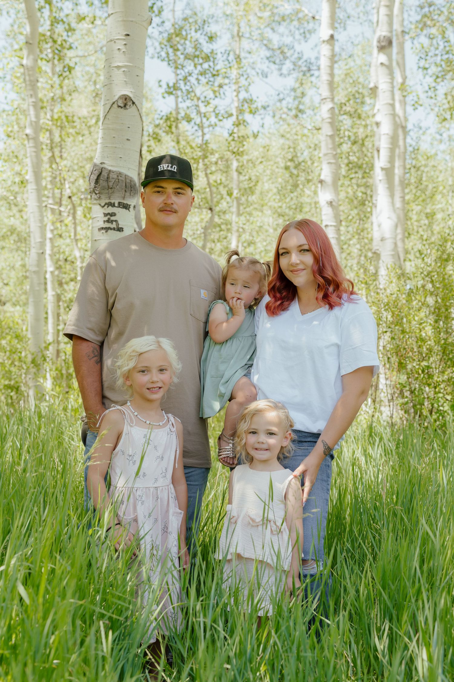 Family of five posing outdoors among tall grass and white birch trees on a sunny day.