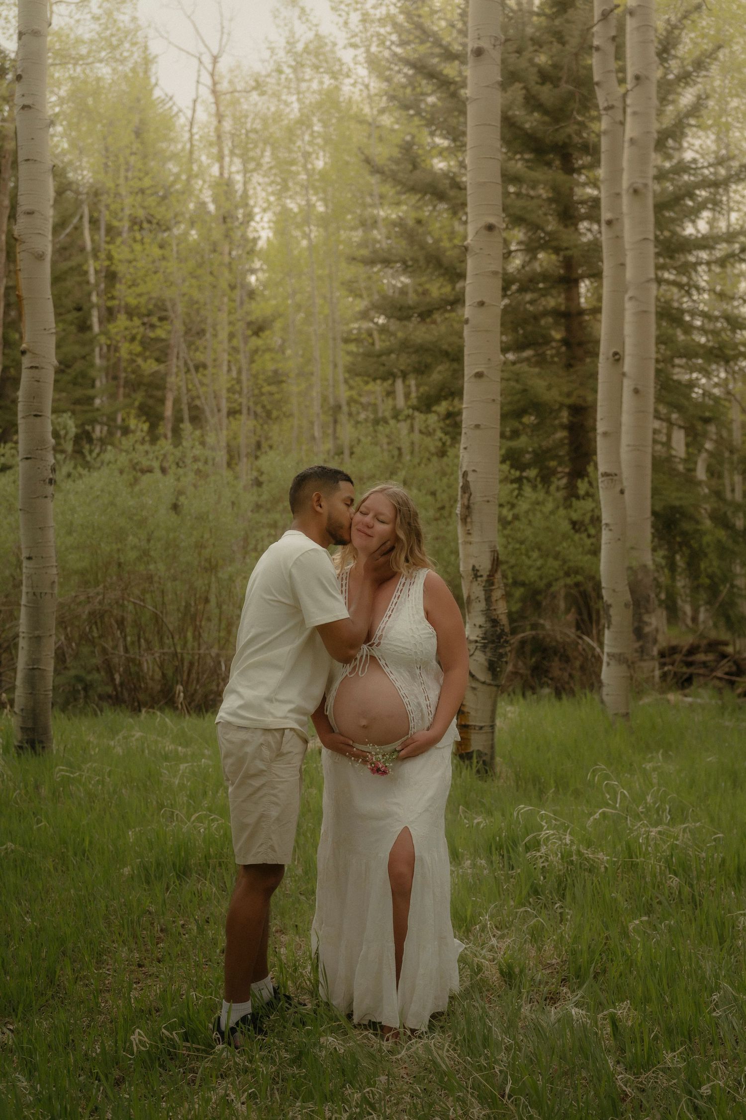 Couple sharing a tender kiss in an aspen forest during a maternity photoshoot, woman in white dress cradling baby bump.