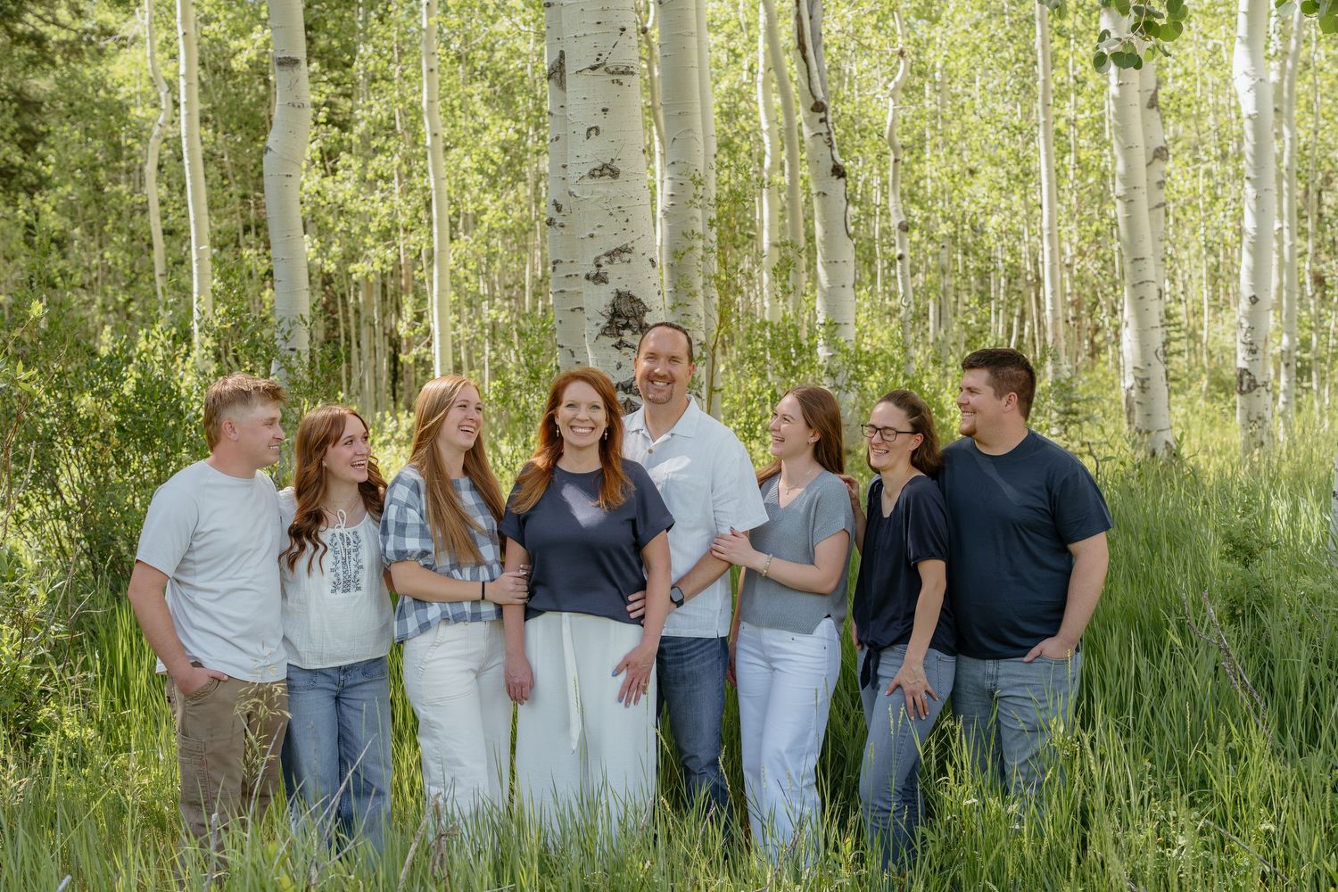 Family of seven smiling together in a lush aspen forest, wearing casual outfits in a bright outdoor setting.