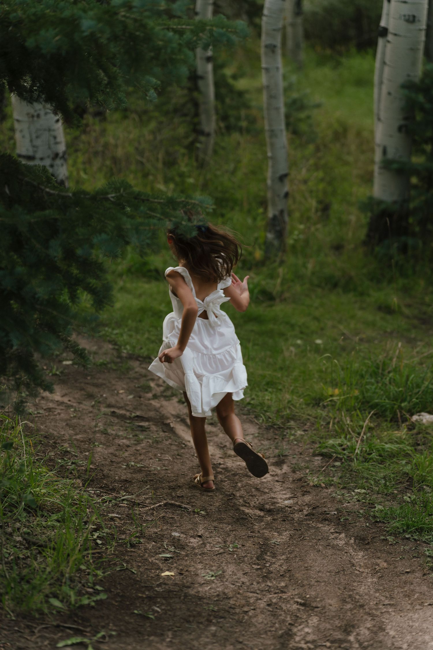 A young girl in a white dress runs barefoot along a dirt path through a lush green forest with aspen trees.