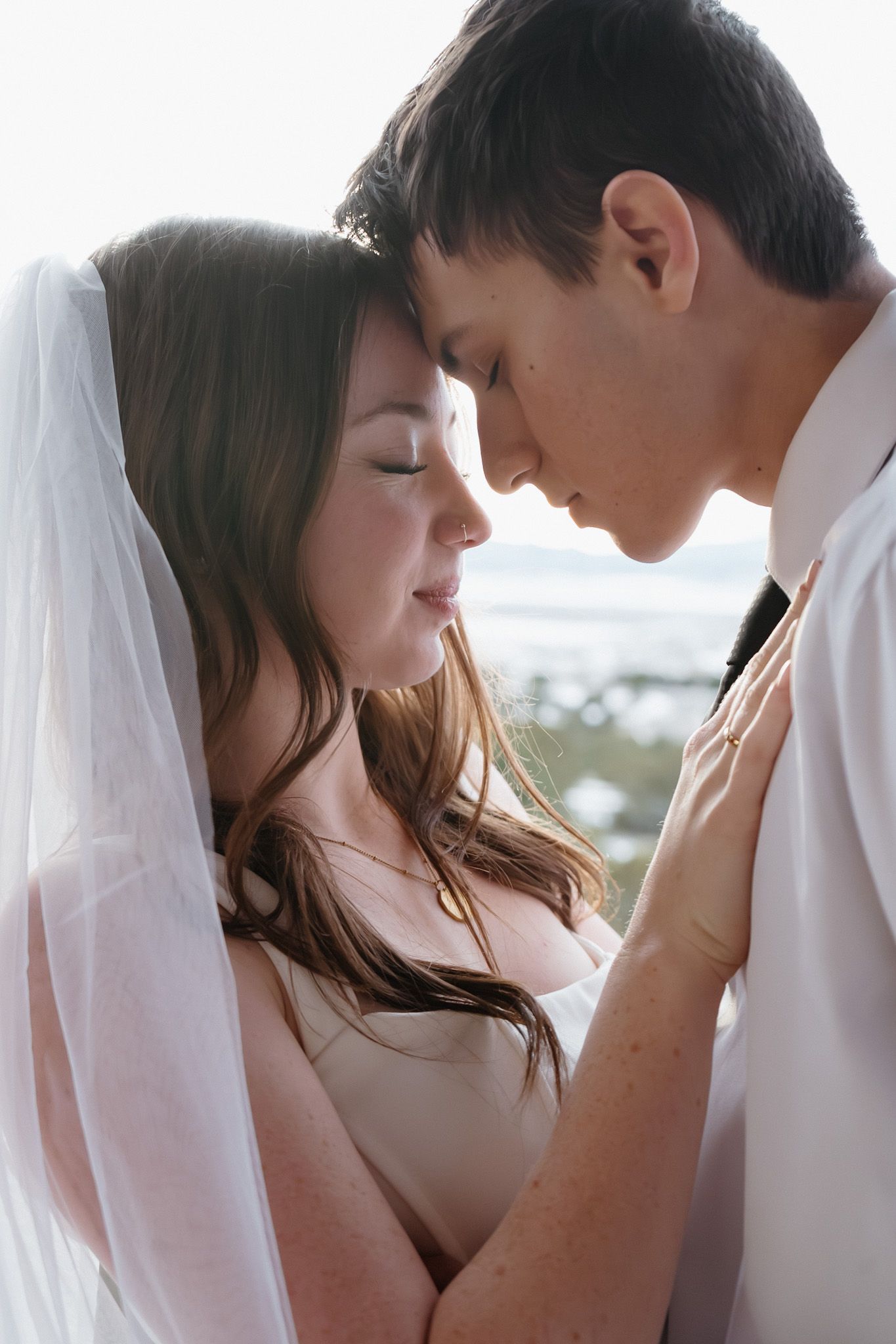 Romantic close-up of a bride and groom touching foreheads under a flowing white veil near a bright window.