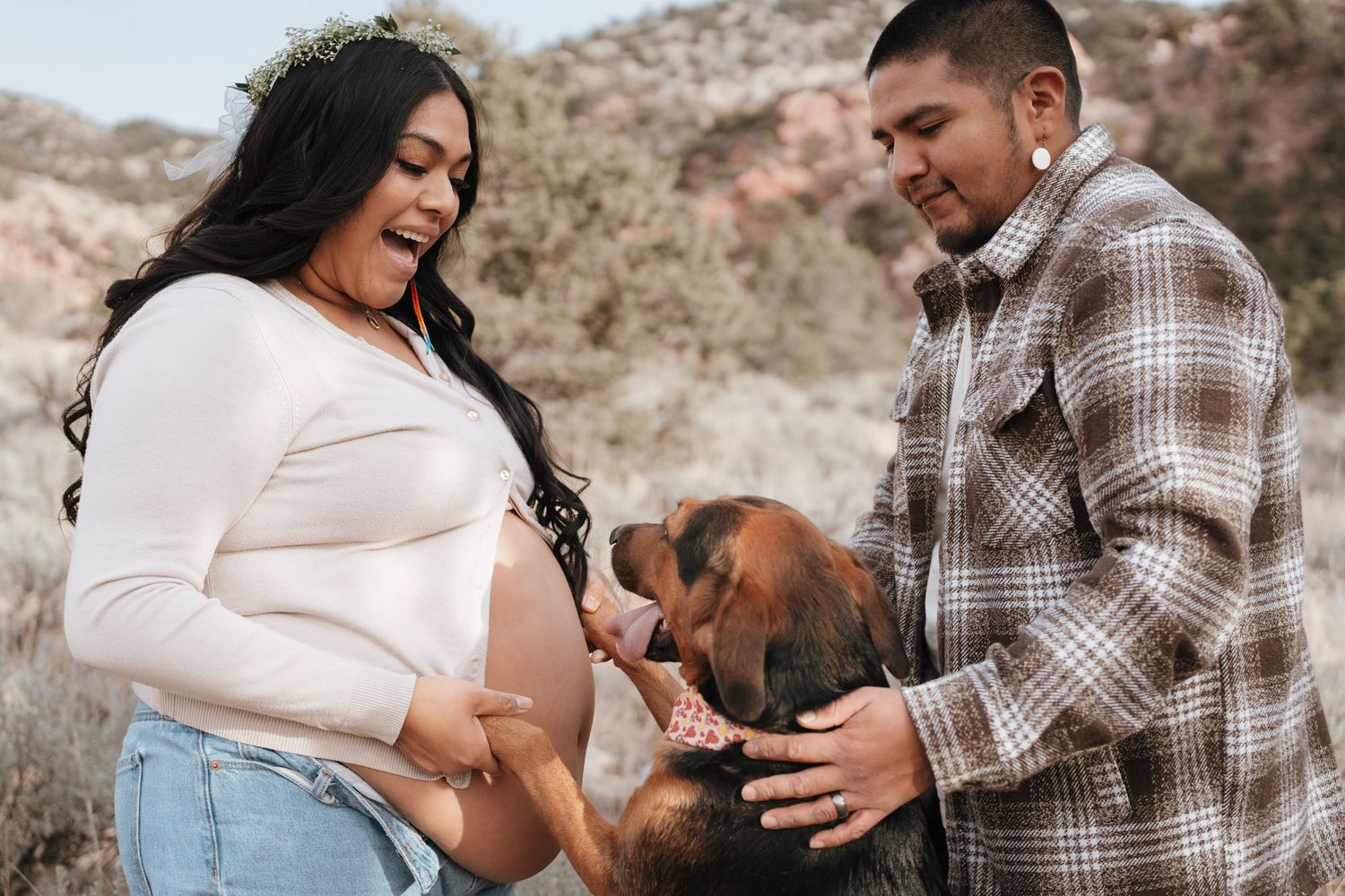 Pregnant couple outdoors with their dog, man in plaid shirt, woman in white top and jeans, desert landscape background.