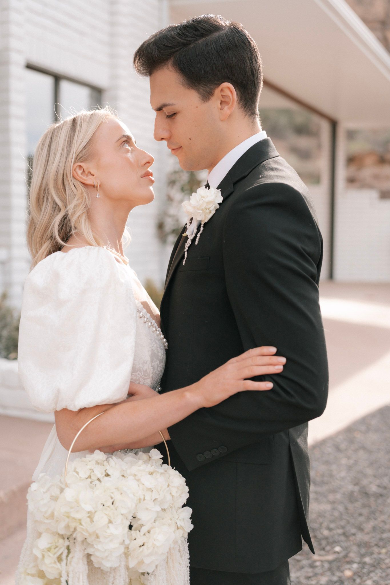 Bride in a white puff-sleeve dress and groom in a black suit share a romantic moment outdoors in sunlight.
