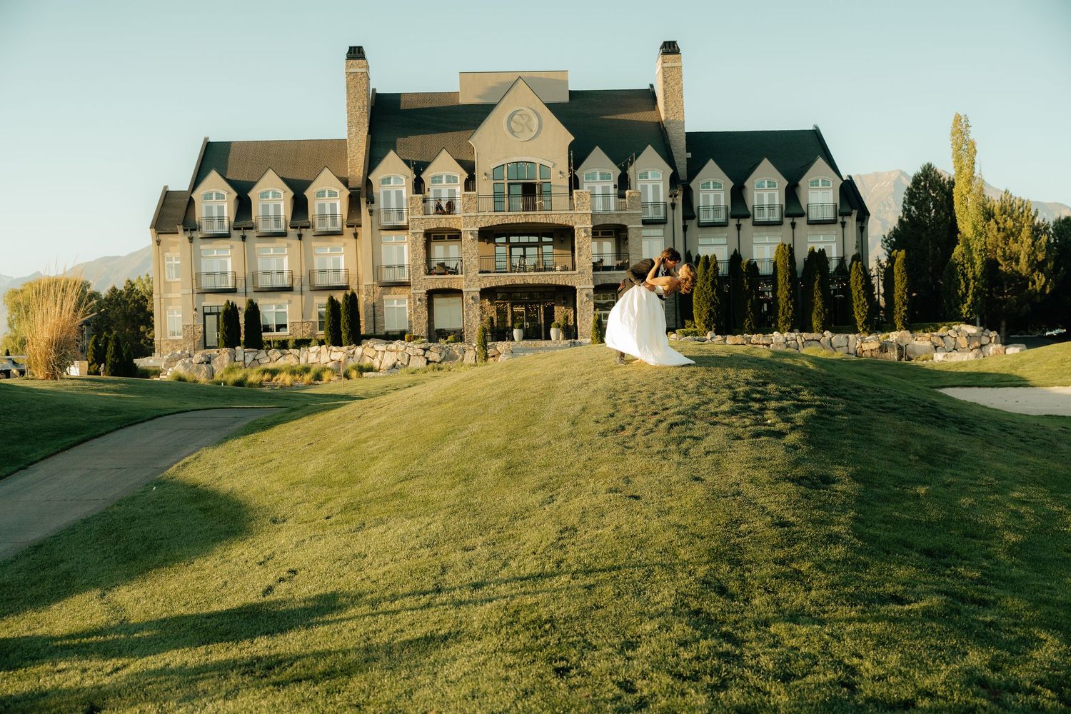Bride and groom embrace on lush green lawn before grand stone mansion wedding venue at golden hour.