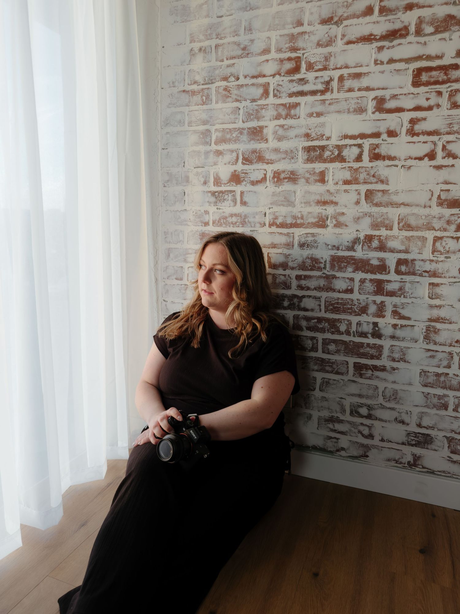 Woman in black outfit holding camera, sitting against rustic brick wall near sheer white curtain in natural light.