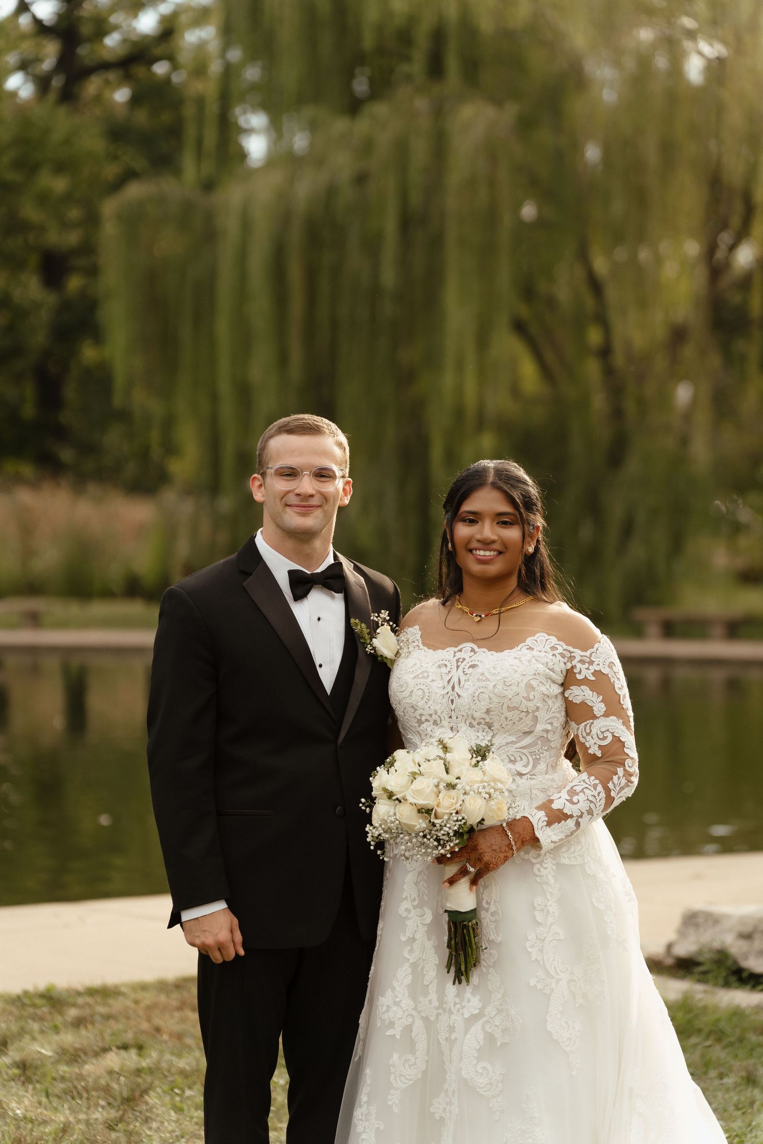 Bride in lace gown holding white bouquet poses with groom in black tuxedo by a pond with willow trees.