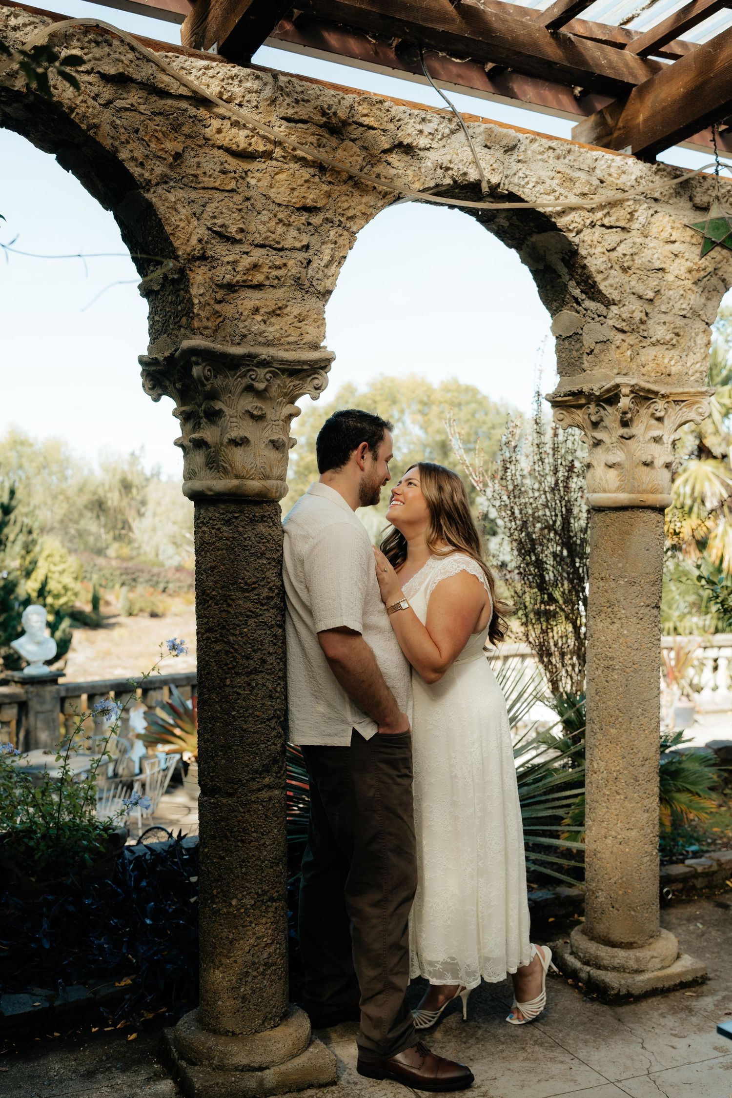 Couple embracing under stone arches in a romantic garden setting, woman in white dress, man in casual attire.