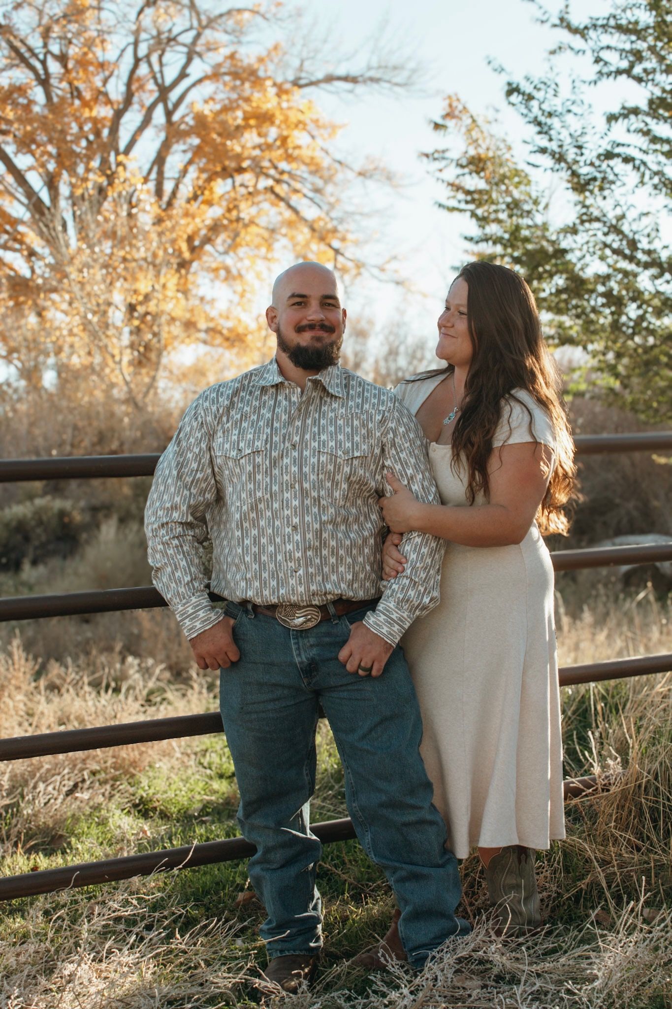 Bald man in western shirt and woman in cream dress posing by rustic fence in autumn setting.
