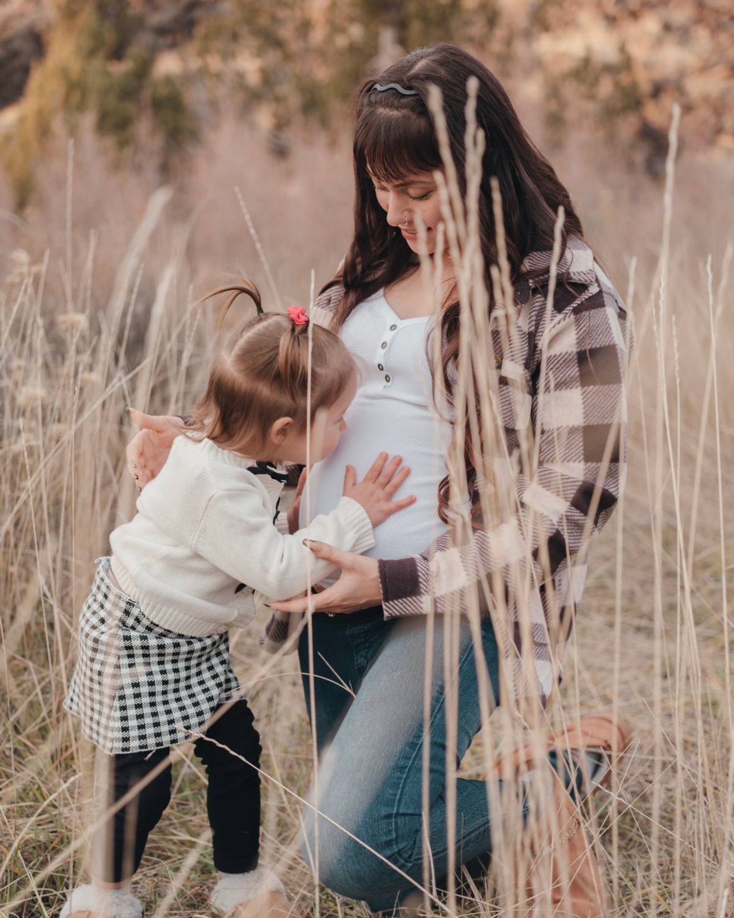 Pregnant woman kneeling in a field while a toddler girl touches her baby bump during an outdoor maternity photo session.