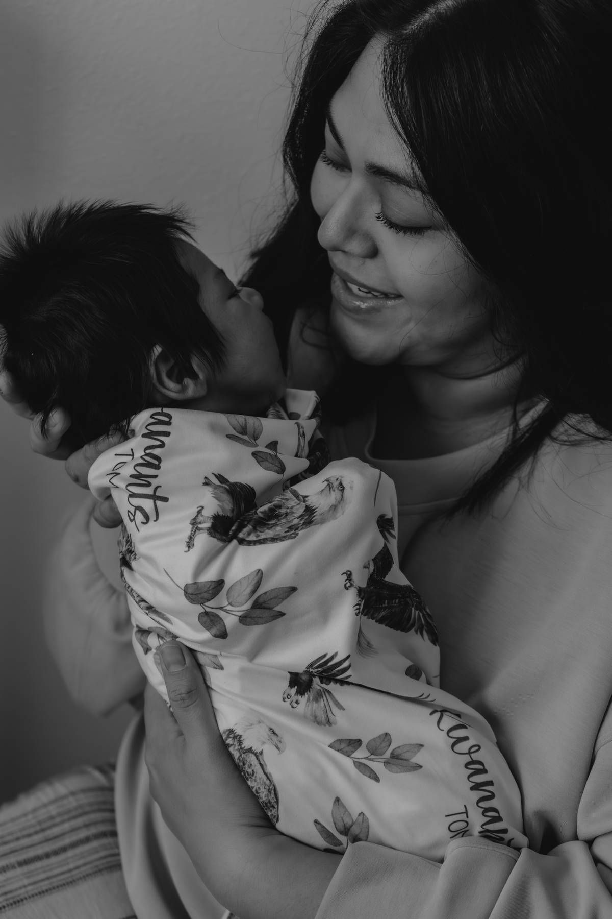 Black and white photo of a smiling mother tenderly holding her newborn baby wrapped in a floral swaddle blanket.