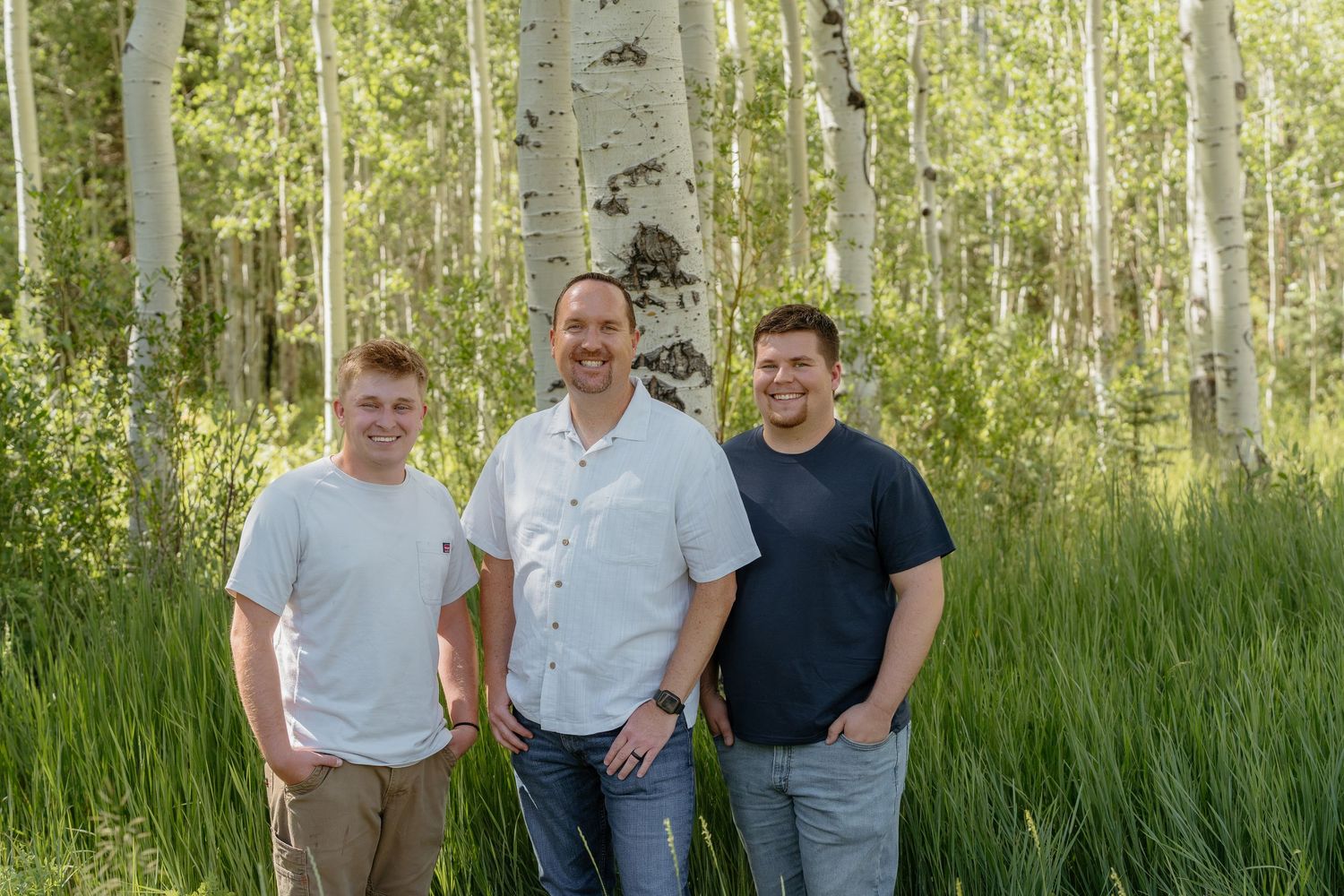 Three men smiling outdoors in a lush aspen forest with white birch trees and green grass.