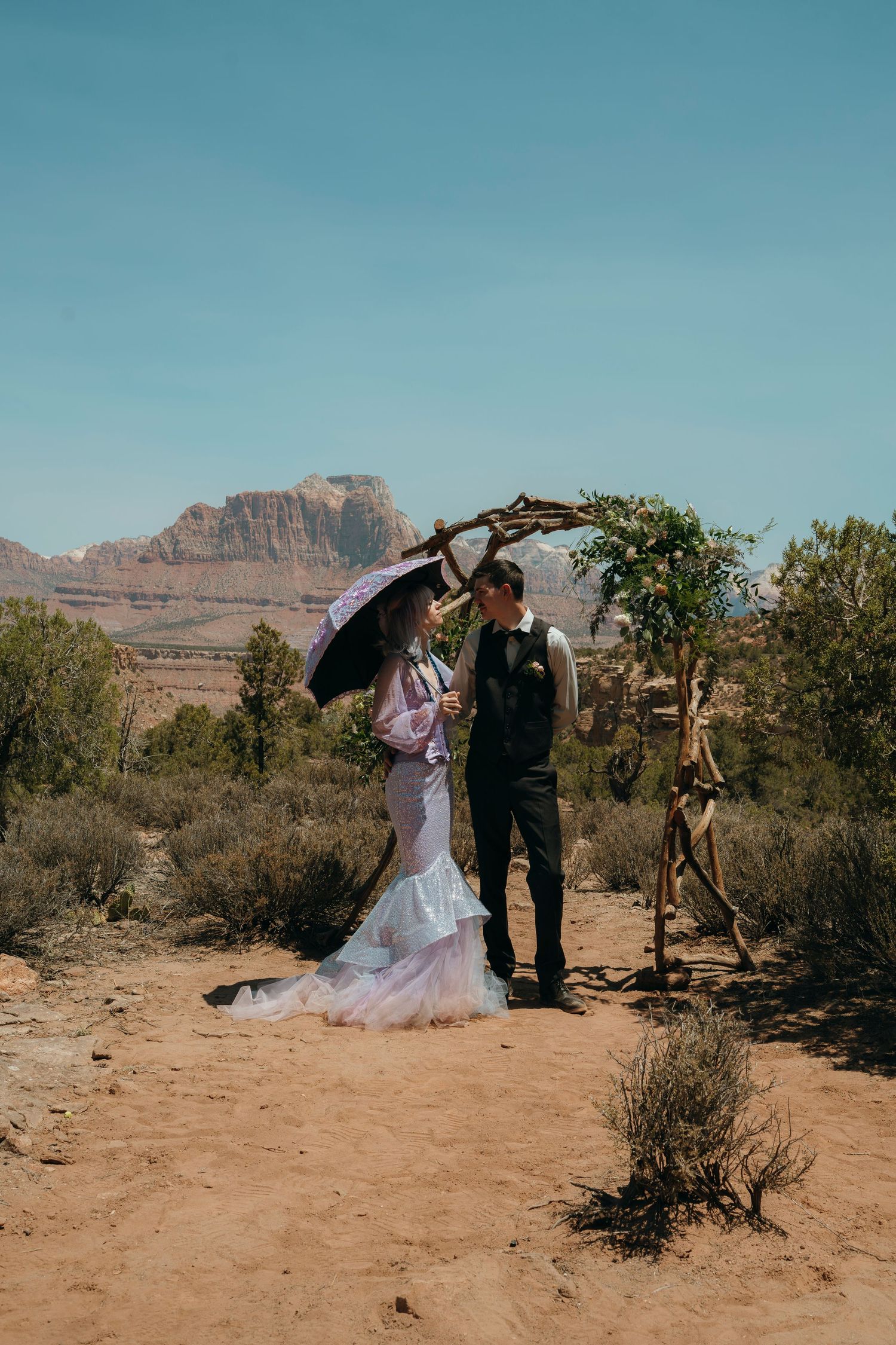 Bride and groom stand under rustic arch in desert landscape with red rock formations during outdoor wedding ceremony.