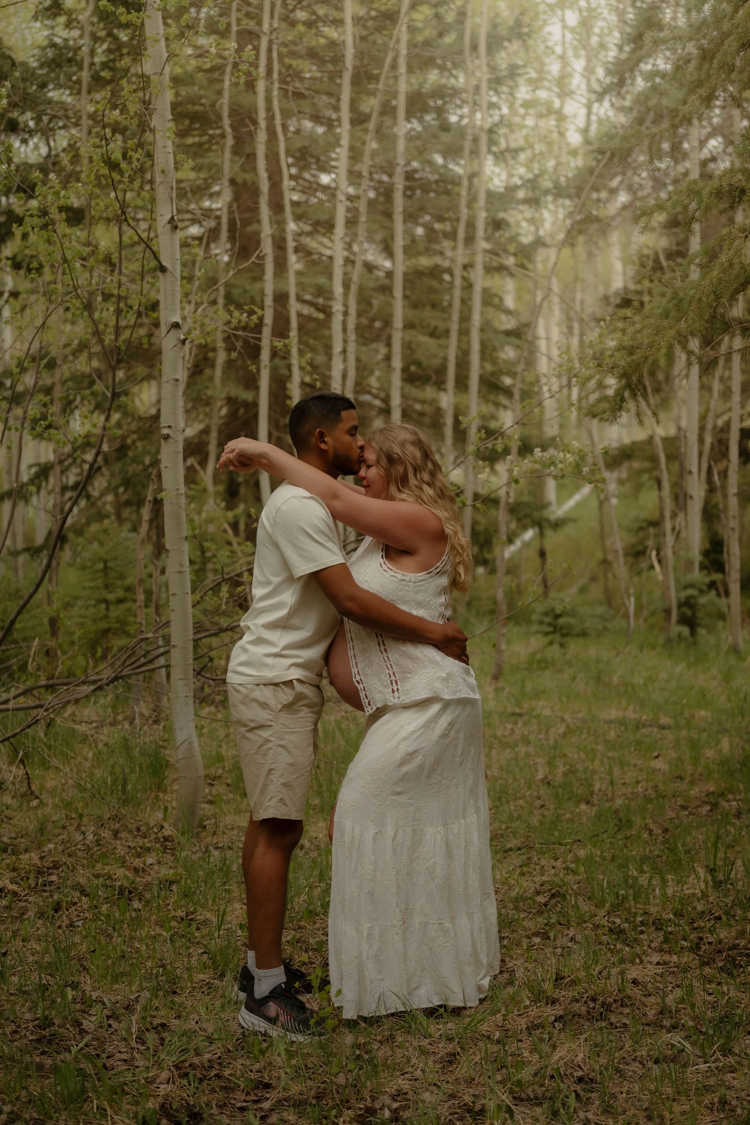 A couple embraces romantically among tall aspen trees in a lush green forest, wearing coordinating white outfits.